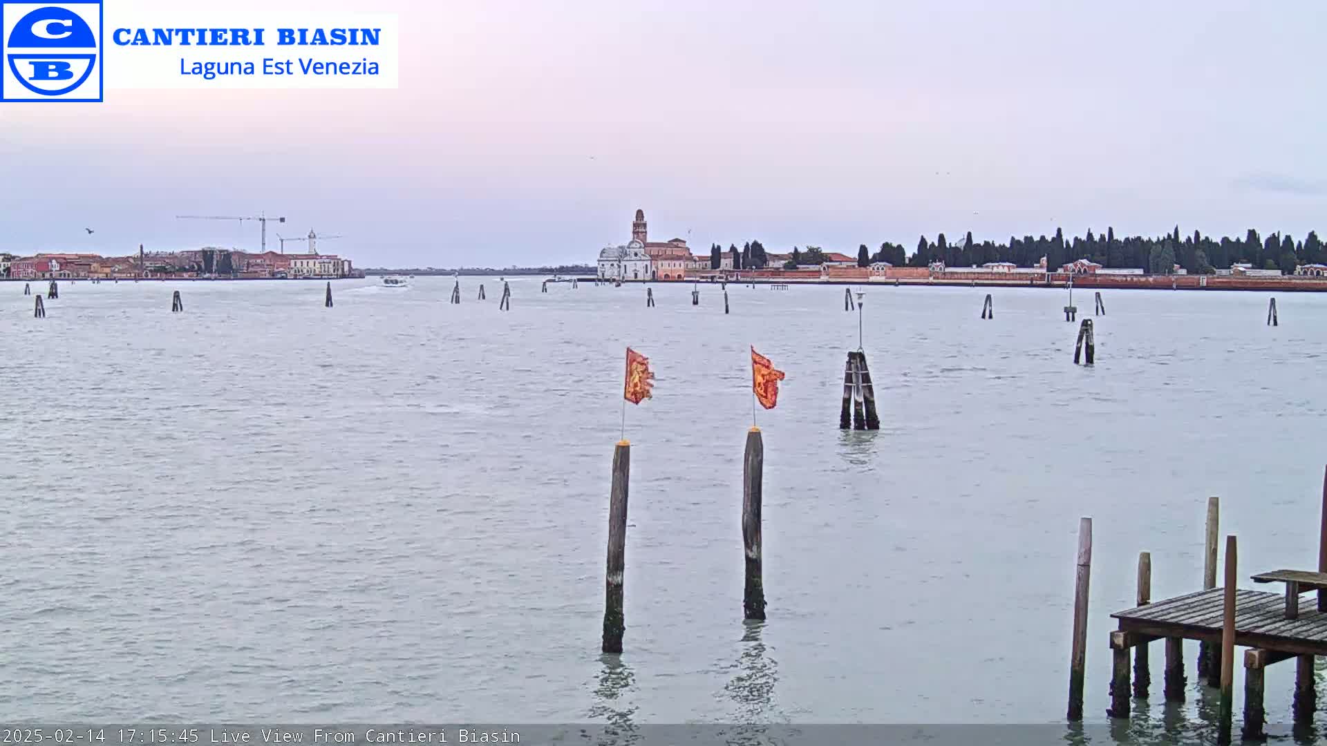 A calm body of water shows a distant cityscape under an overcast sky, with two orange flags on posts in the foreground and a small wooden dock to the right.