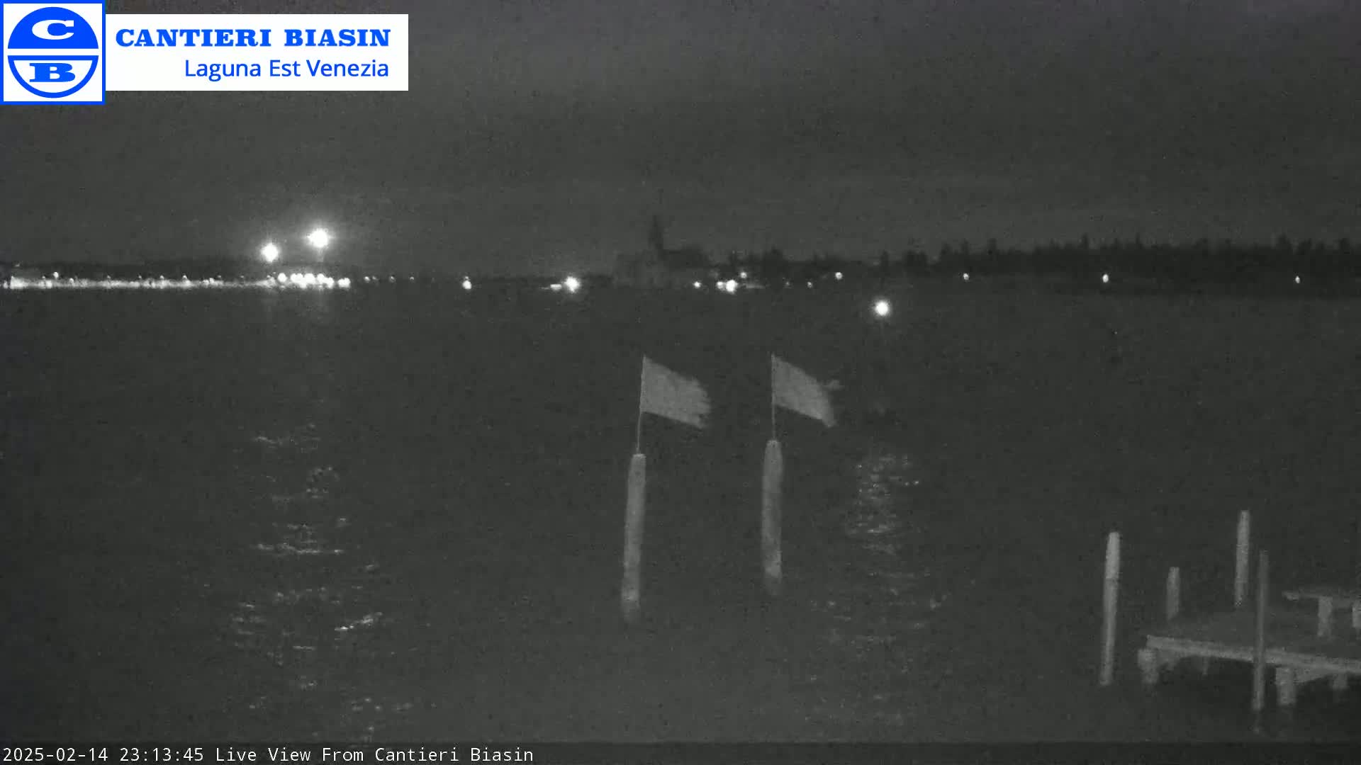 A nighttime, low-light view of a dark body of water with two flags on posts and a small dock in the foreground, and lights visible on the opposite shore.