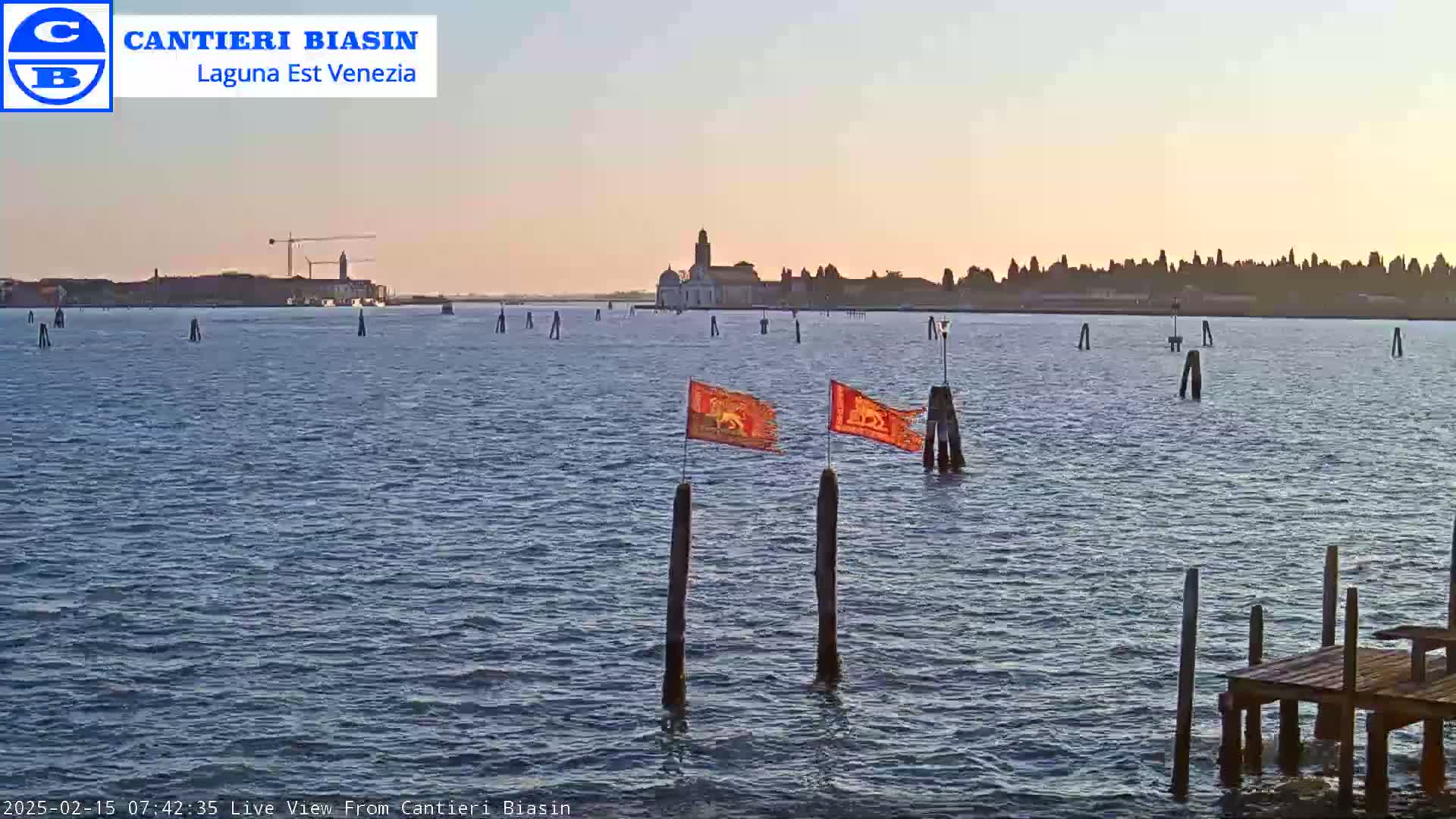 Two orange flags fly from wooden posts in a calm body of water, with a distant cityscape and a construction crane visible under a clear, twilight sky.