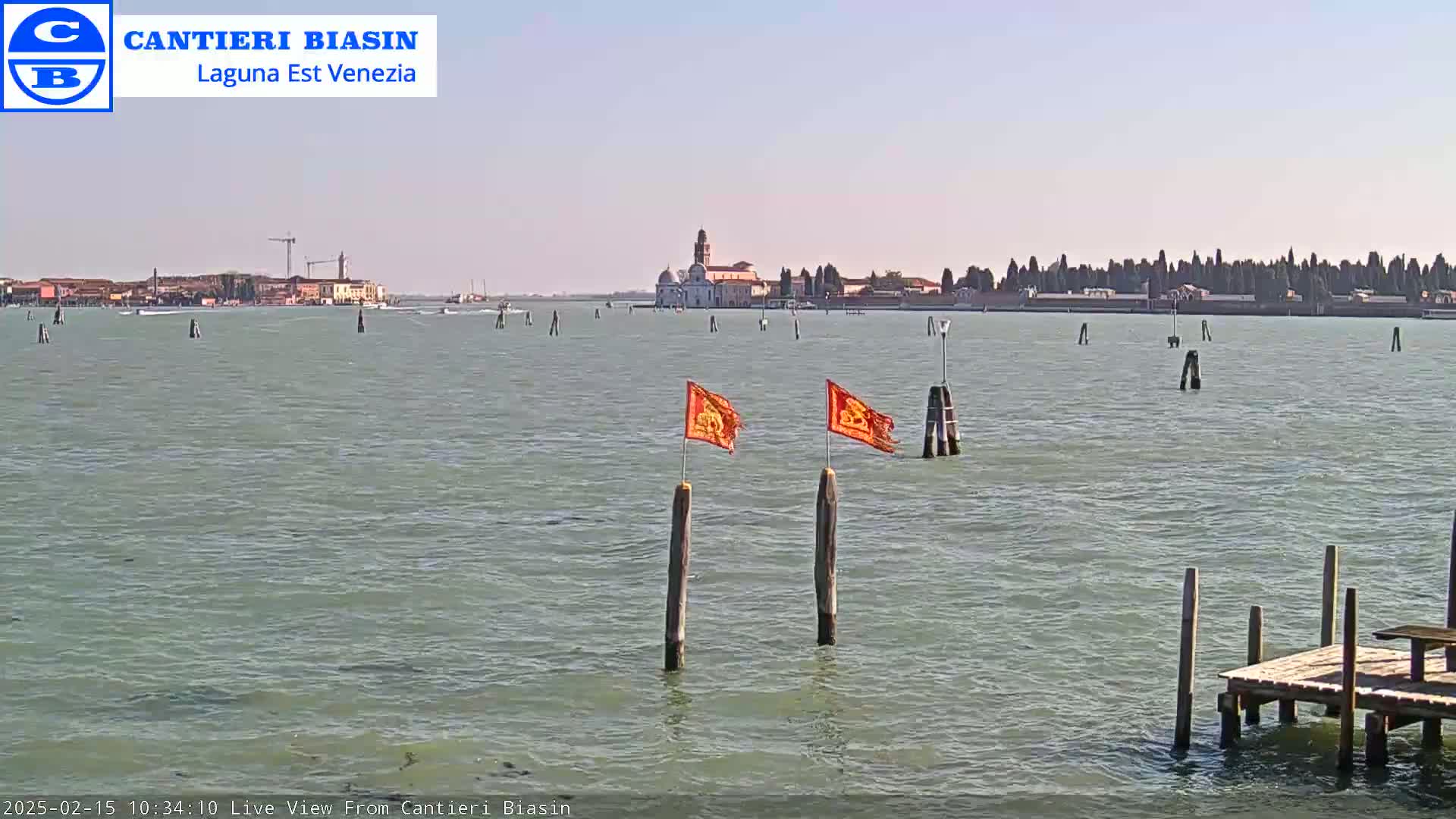 A calm body of water shows a distant island with buildings and trees under a clear sky, with two orange flags on wooden posts in the foreground.
