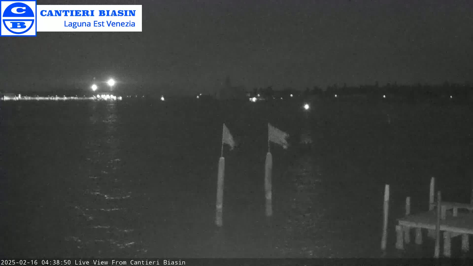 A nighttime, low-light view of a calm body of water with two flags on poles in the foreground and a distant shoreline faintly visible.