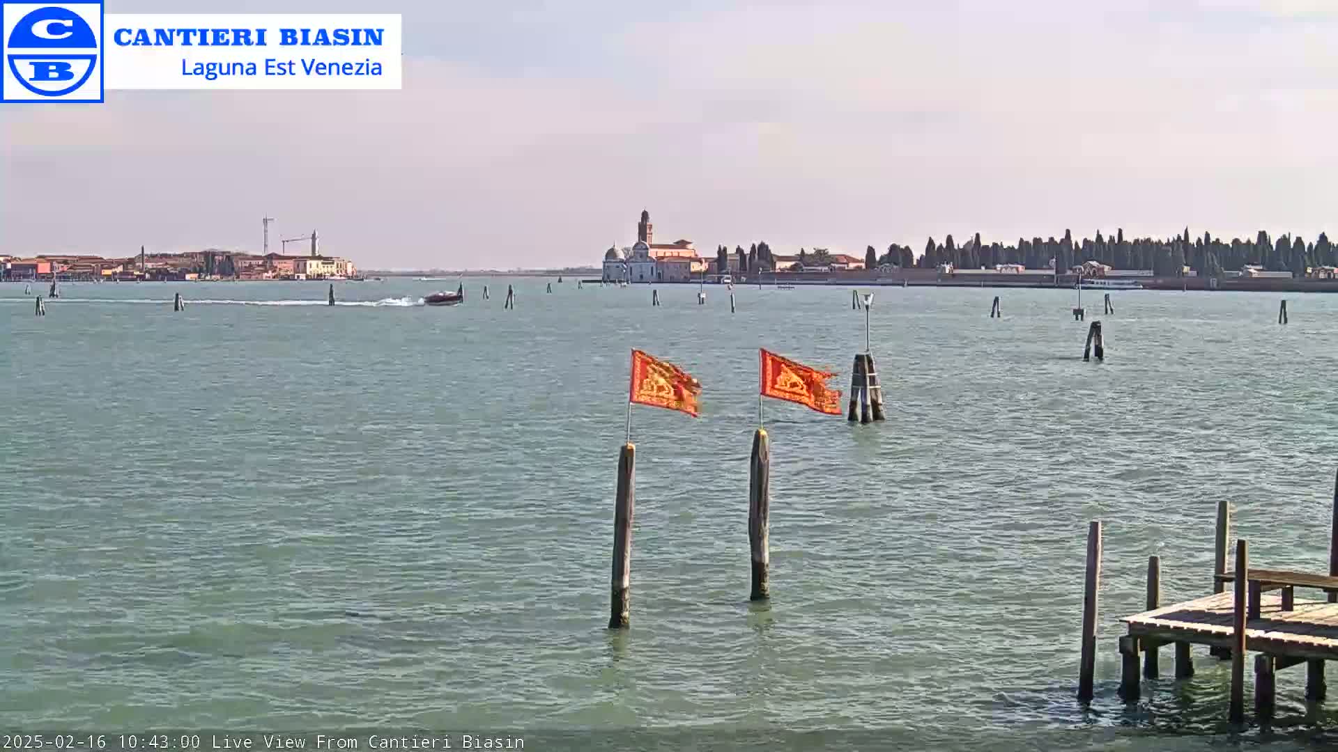 A calm body of water shows a distant island with buildings, a small boat, and two orange flags on wooden posts in the foreground, under a clear sky.