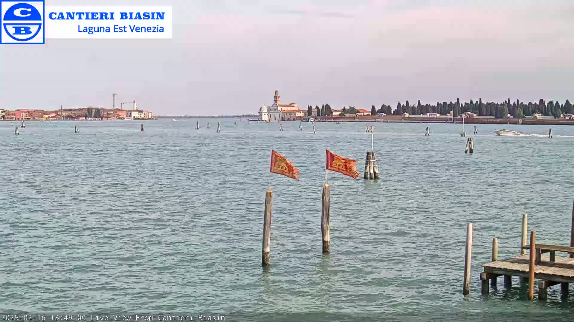 A calm body of water shows a distant island with buildings and trees under a pale sky, two tattered orange flags on posts in the foreground, and a small dock to the lower right.