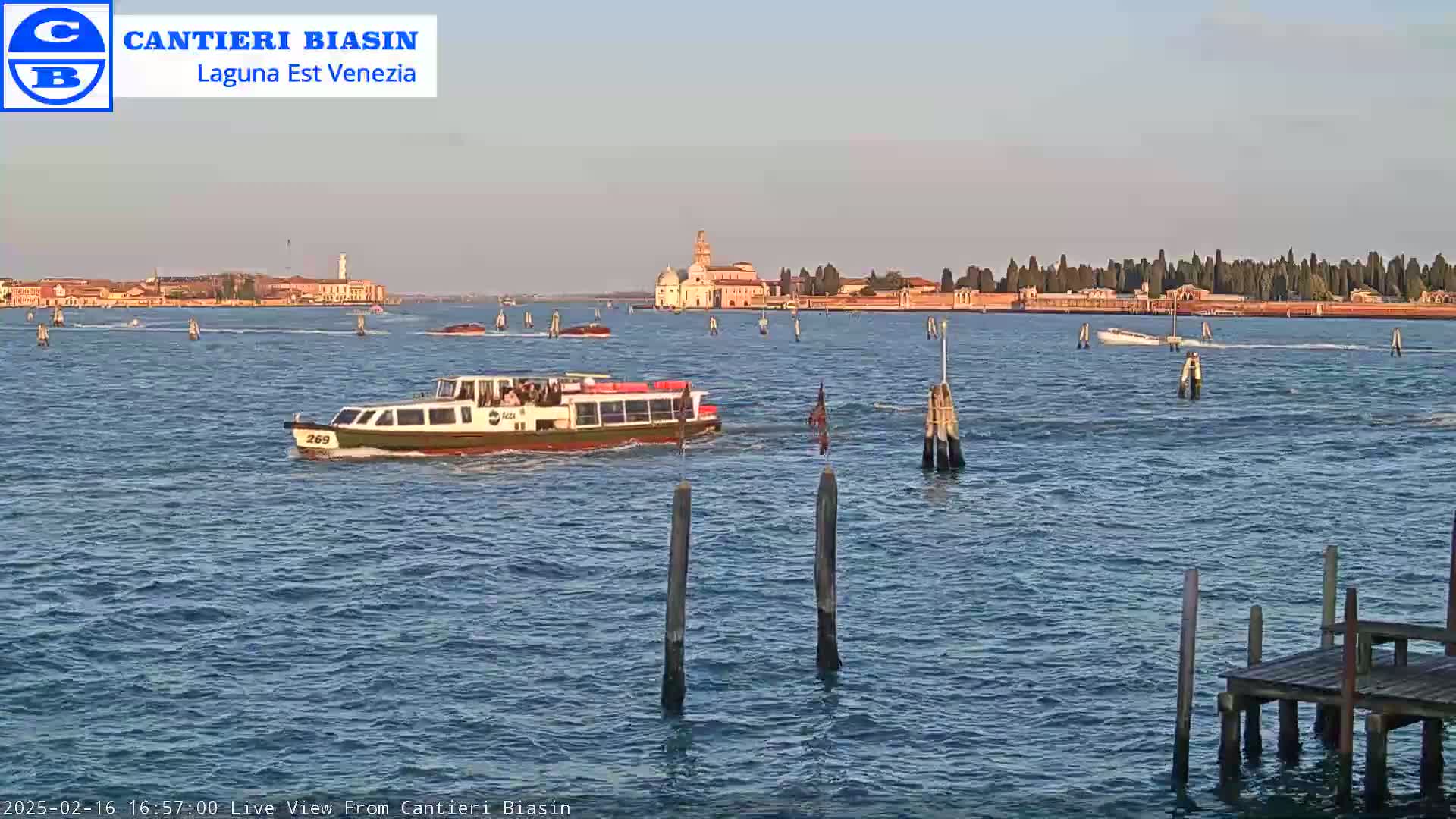 A passenger boat travels on calm water past wooden pilings and toward a distant island with buildings under a clear, light-blue sky.