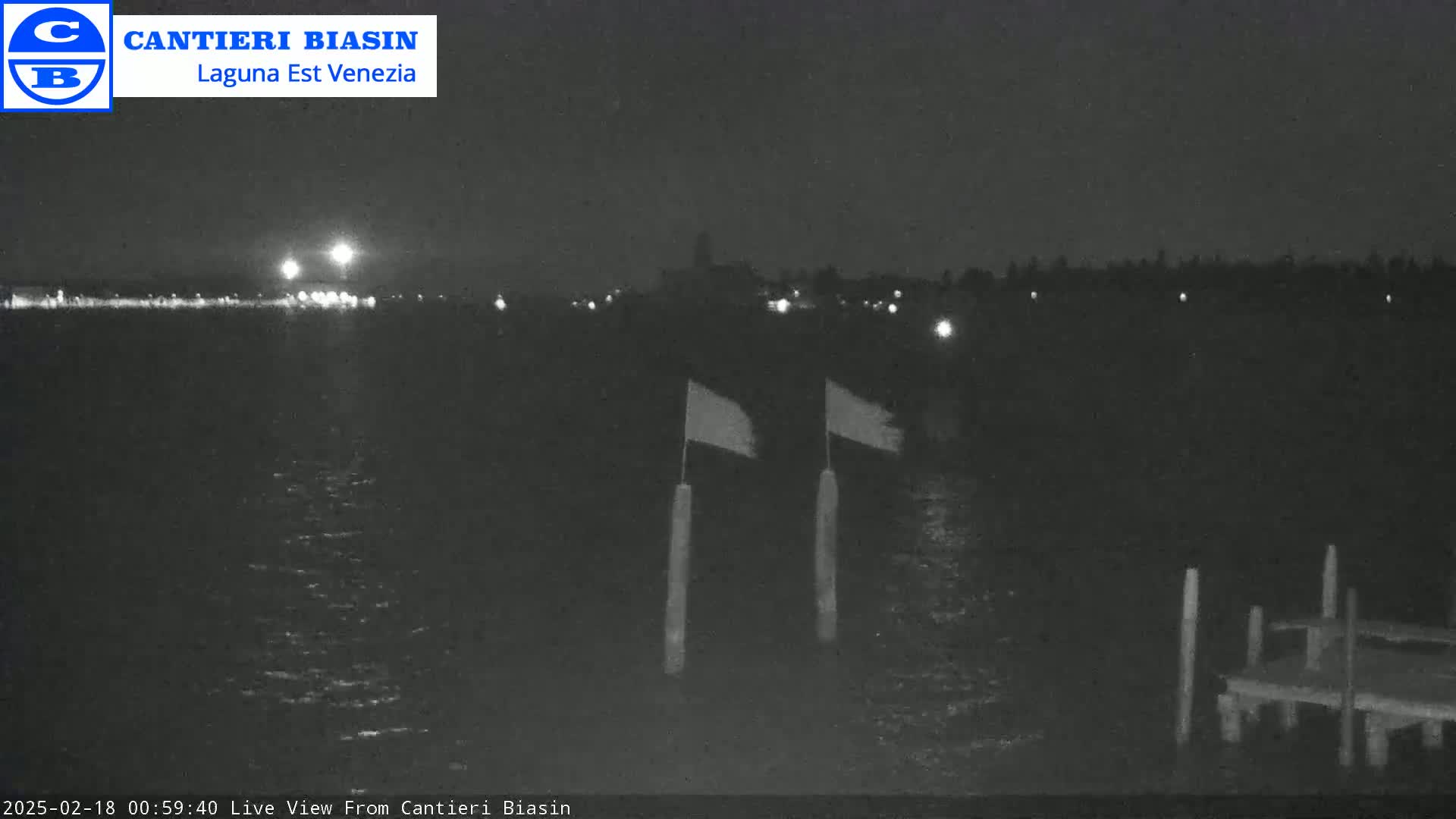 At night, a dark body of water reflects lights from a distant shore, two flags hang on posts near a small dock.