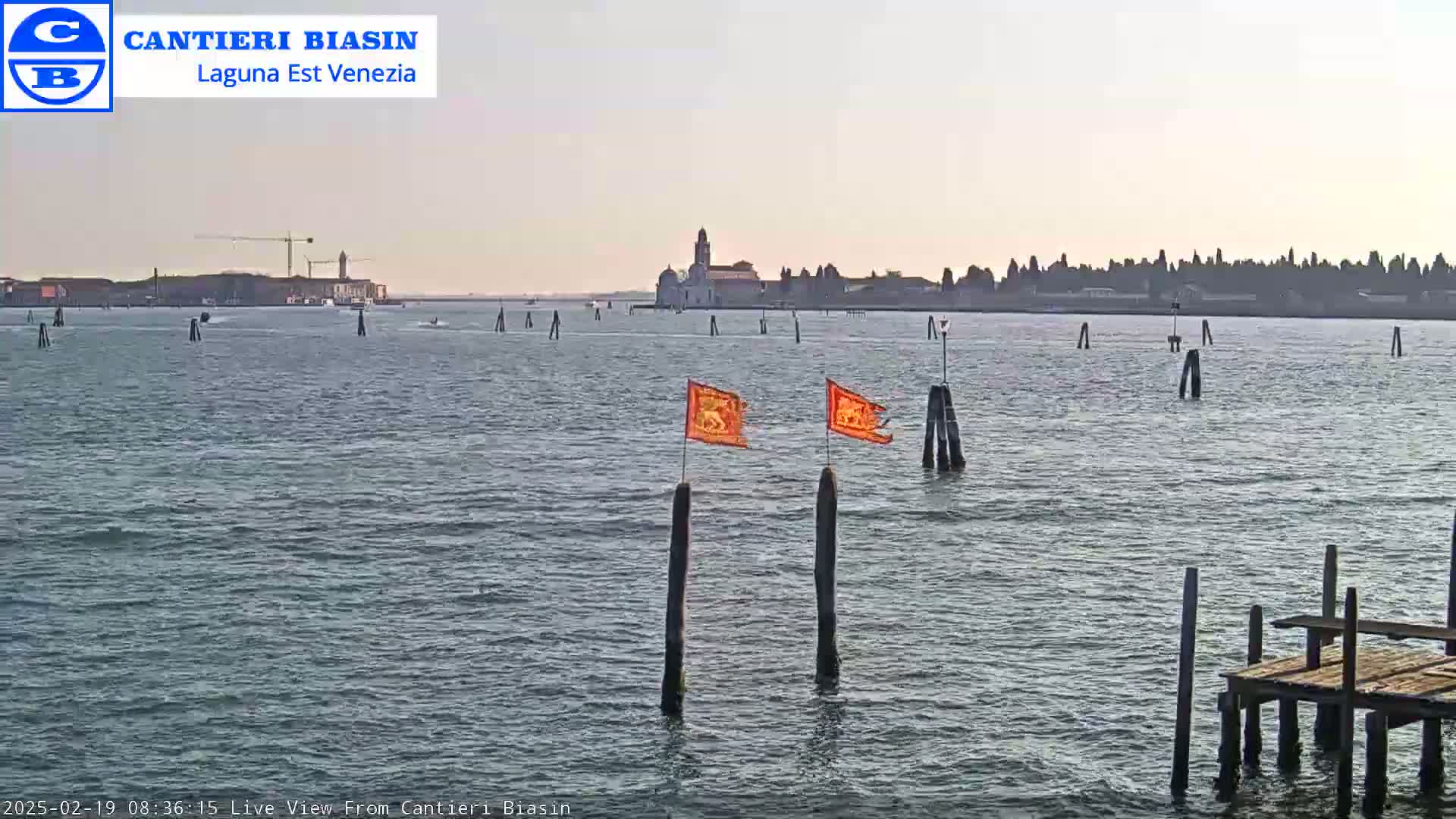 A calm body of water shows a distant island with buildings, a construction crane, and two orange flags on posts in the foreground under a light overcast sky.