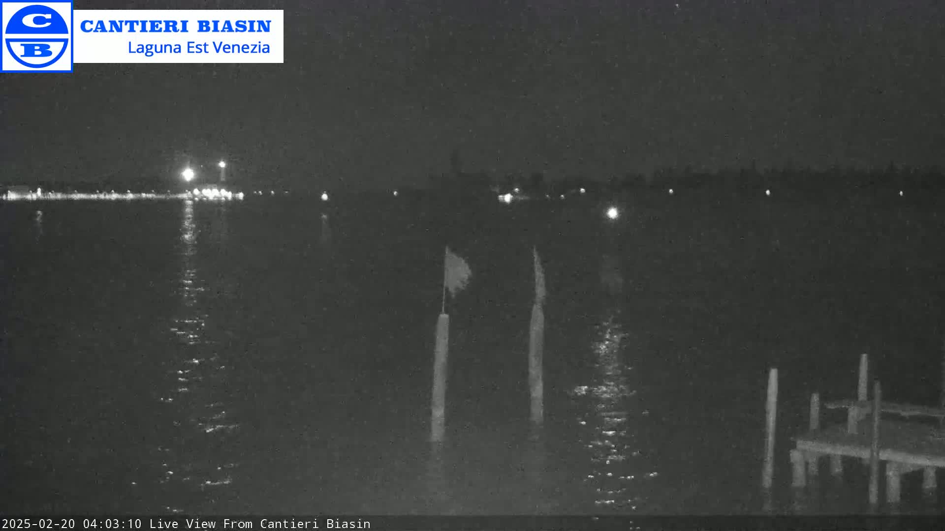 A nighttime view of calm water with a distant shoreline showing lights, and a small dock with posts and flags in the foreground.