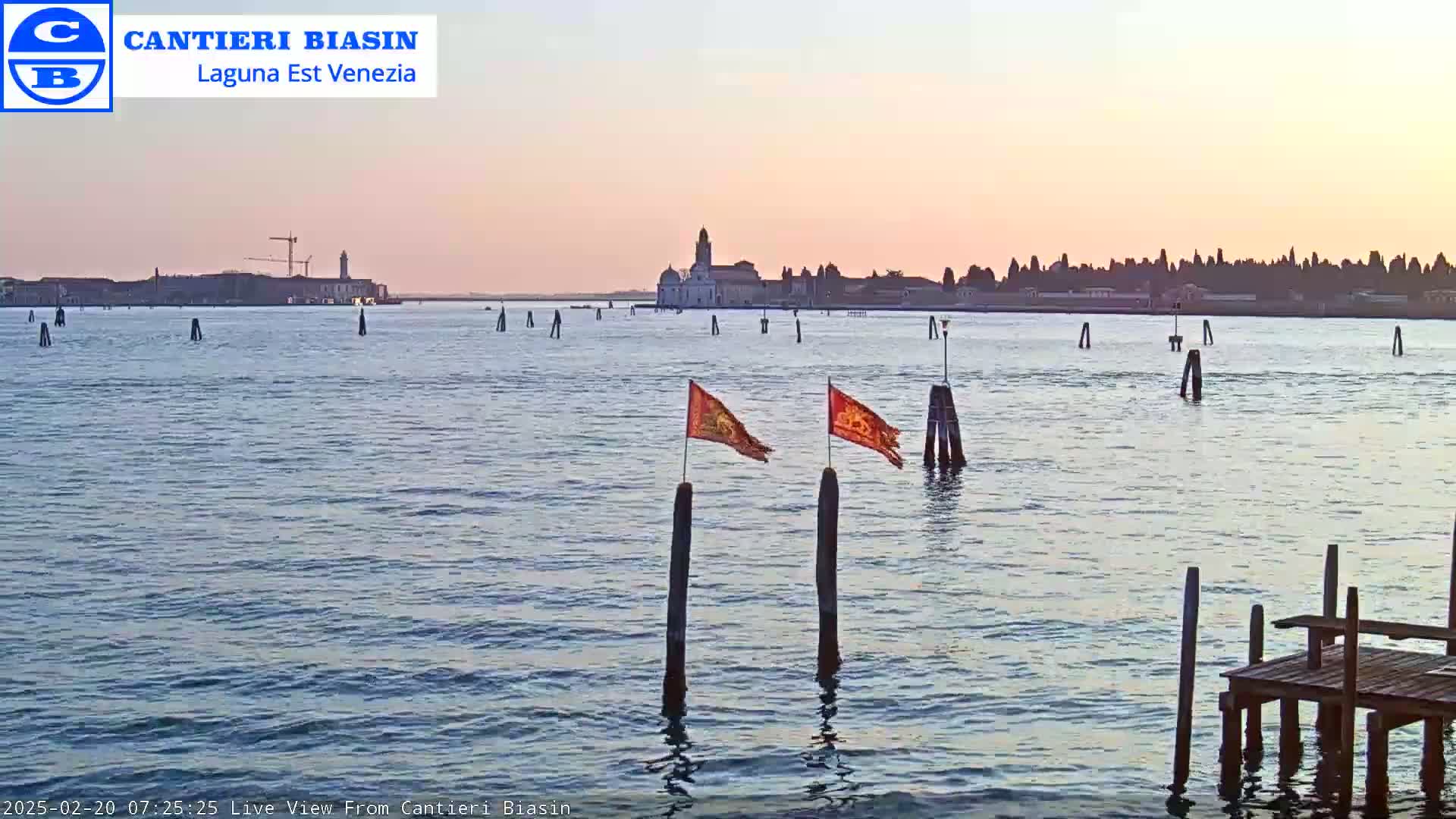 A calm body of water at sunset, with a distant island showing buildings and a bell tower, and two small orange flags on posts in the foreground.