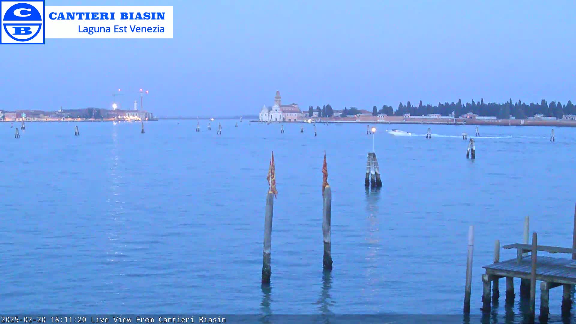 A calm, twilight view across blue water shows a distant island with buildings and trees, a small boat, and two wooden posts with flags in the foreground near a small wooden dock.