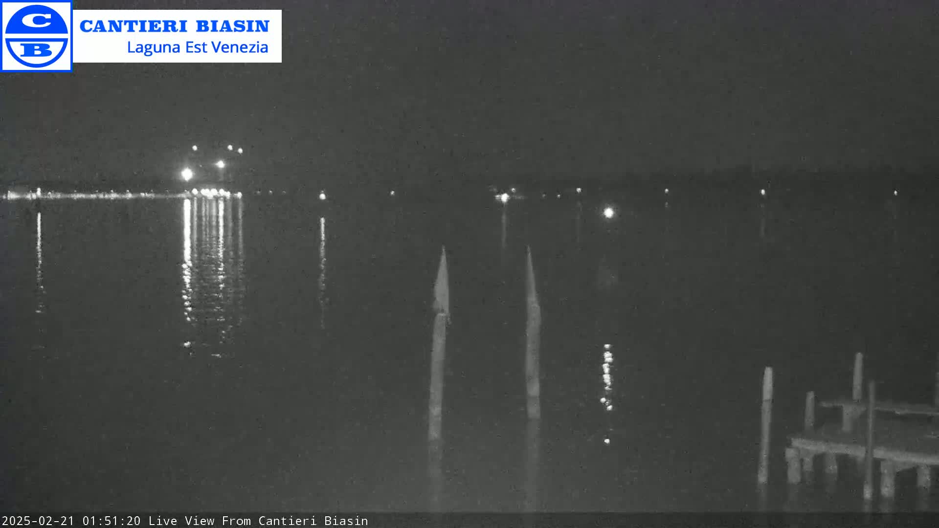 A nighttime view of a dark body of water with lights reflecting on its surface, two tall thin objects in the water, and a dock in the foreground.