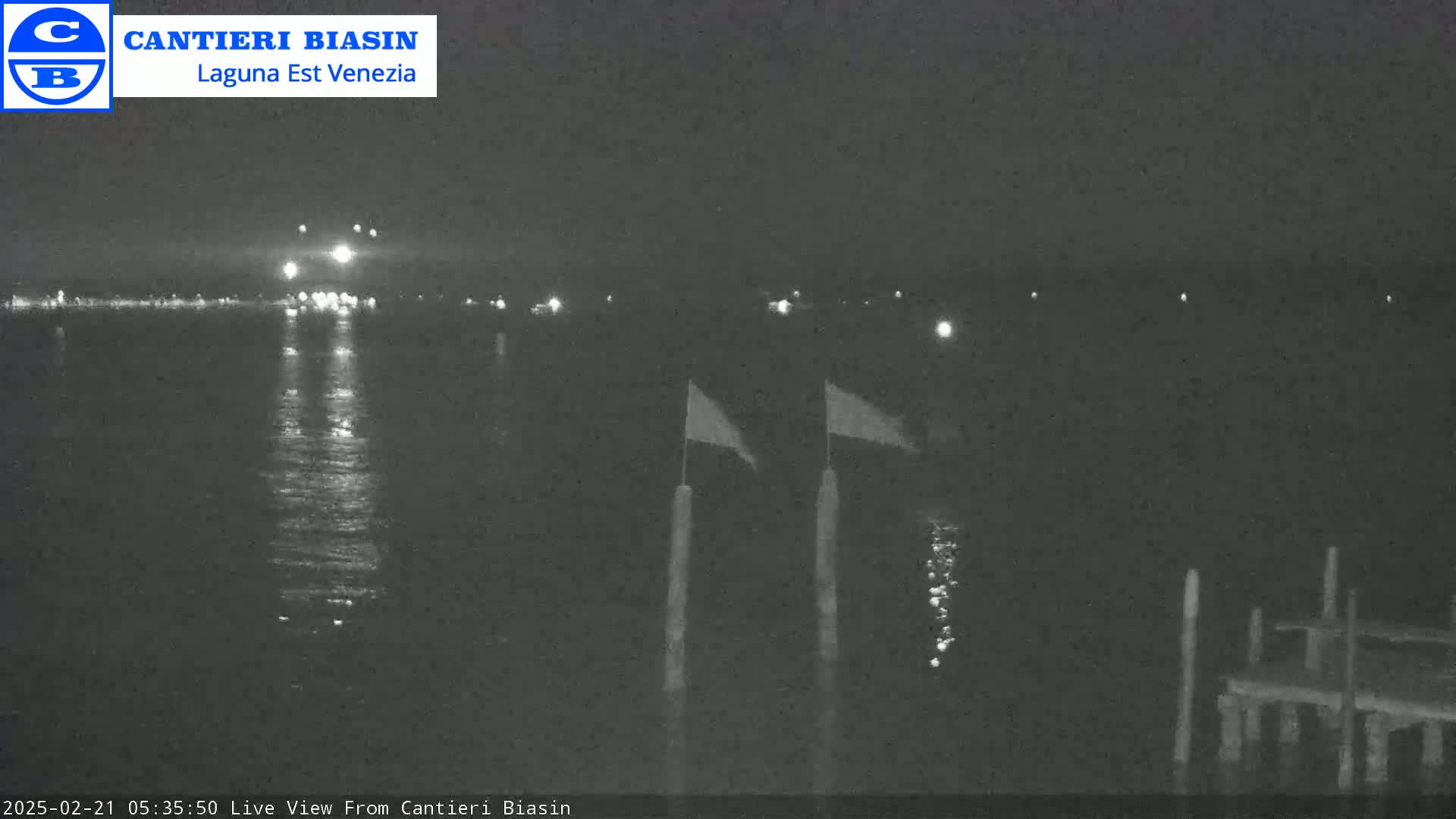 A nighttime view of calm water reflecting lights from a distant shore, with two flags on poles in the foreground and a dock to the right.