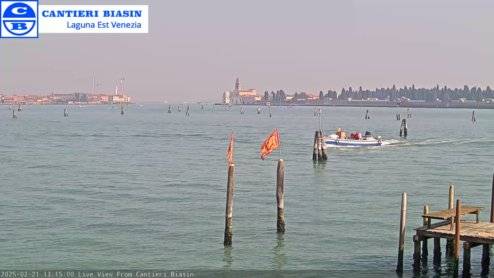 A small boat passes by a calm body of water with wooden posts and flags, while a distant island with buildings is visible under a hazy sky.