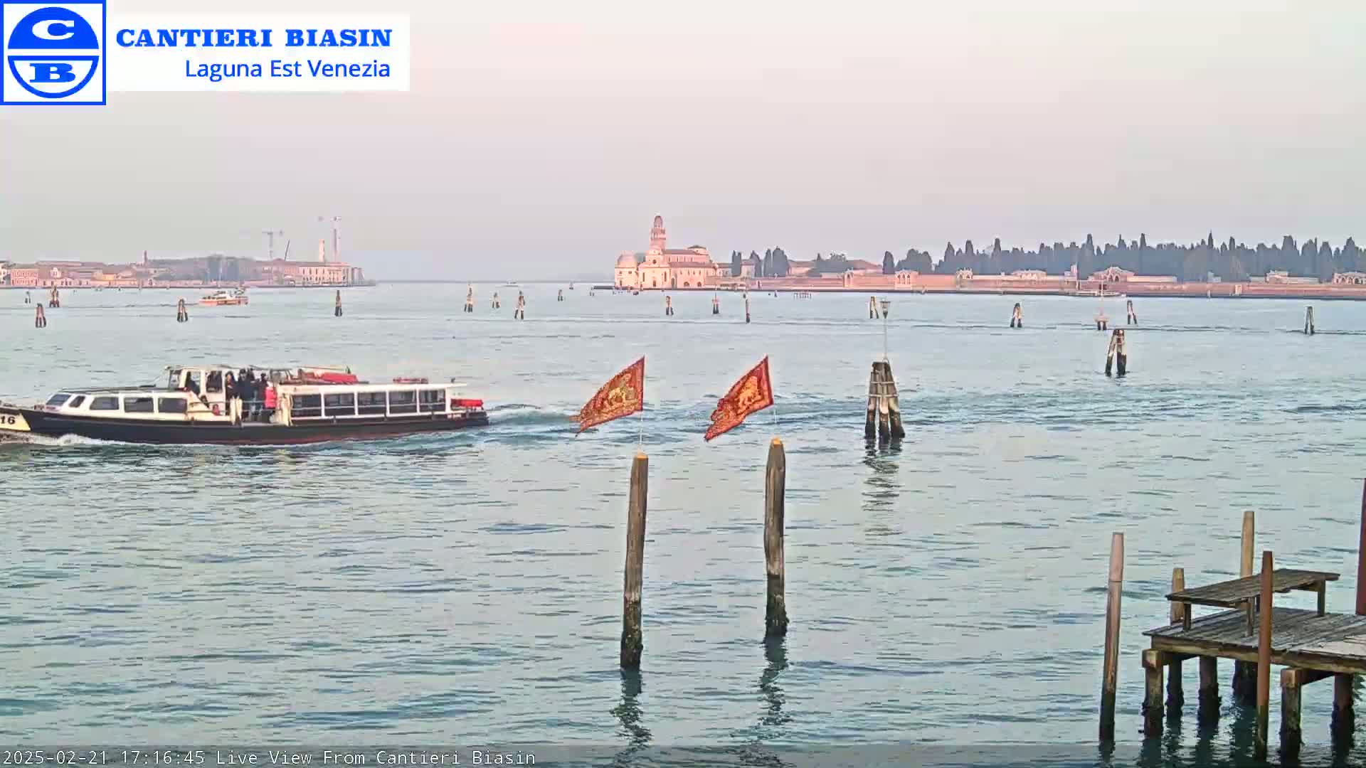 A passenger boat travels on calm, light-grey water toward a distant island with buildings under a hazy sky.