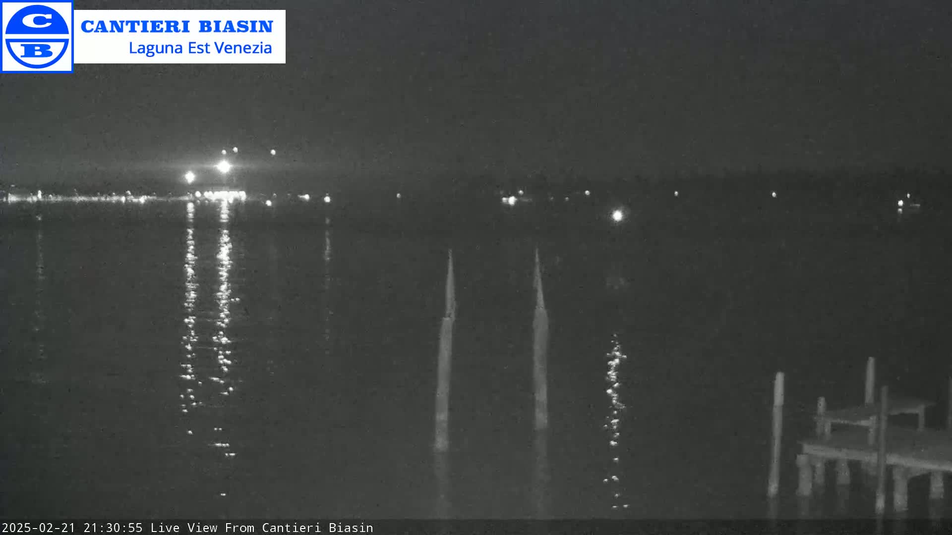 A nighttime view of calm water reflecting lights from a distant shore, with three vertical poles and a small dock visible in the foreground.