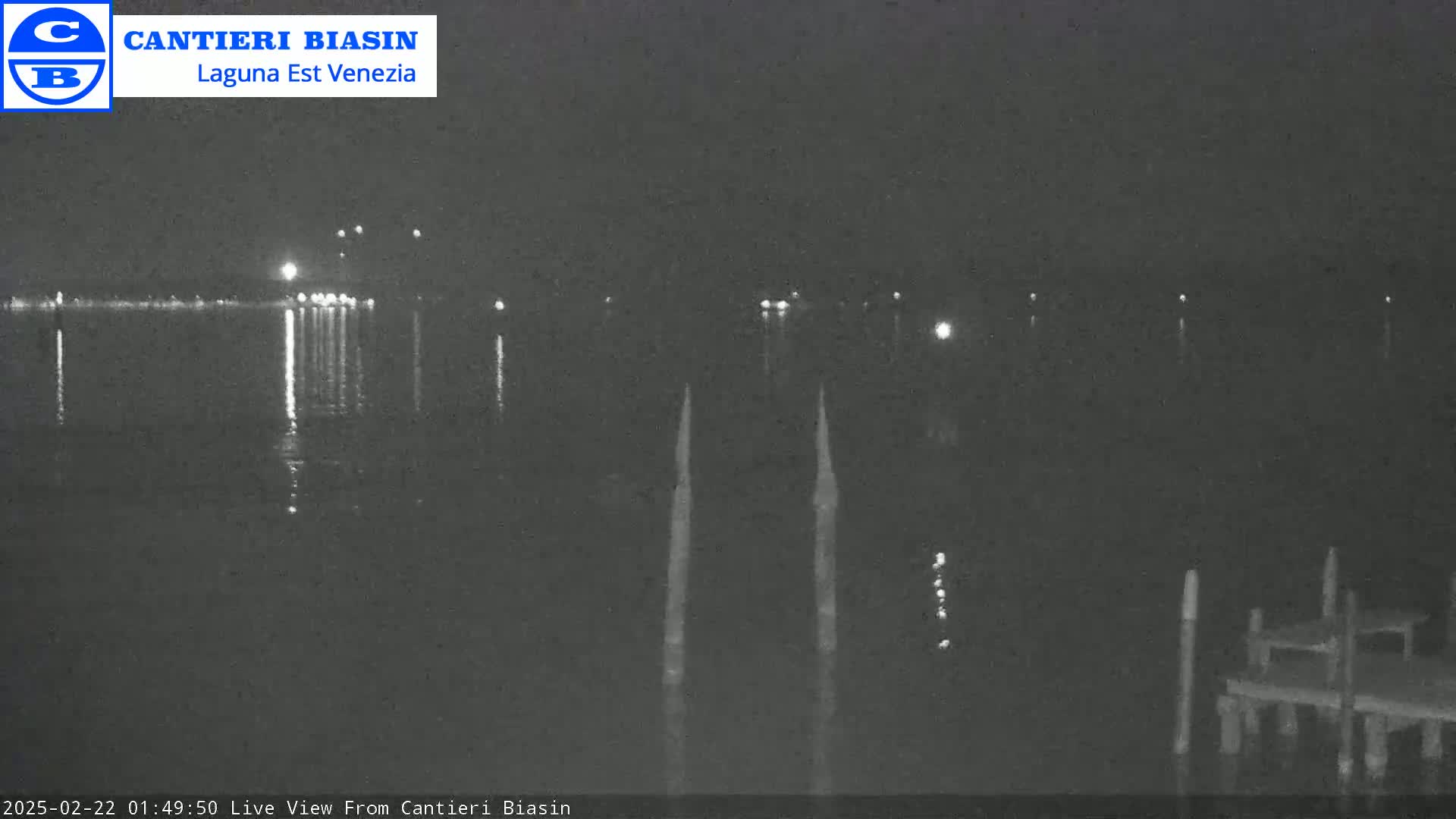 A nighttime, low-light view of a calm body of water shows distant lights on the opposite shore, reflected in the water, with a small dock and pilings in the foreground.