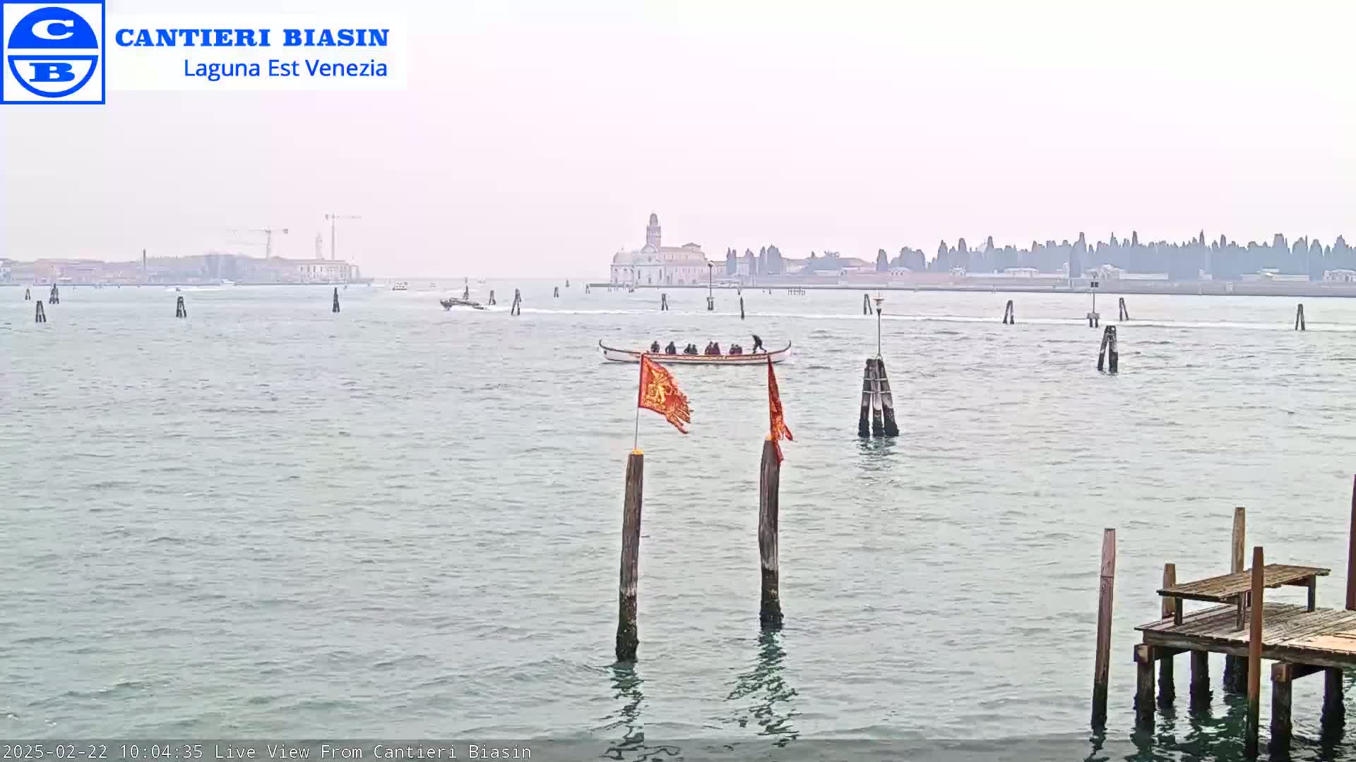 A long shot of a waterway shows a gondola with passengers, a distant city skyline, and several wooden posts in the water under an overcast sky.