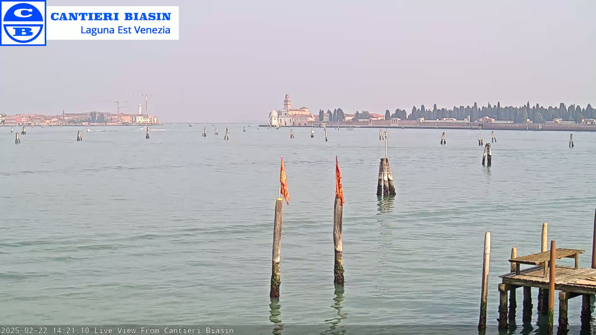 A calm body of water shows a distant island with buildings and trees, and closer to the foreground, wooden posts with orange flags and a small wooden dock.