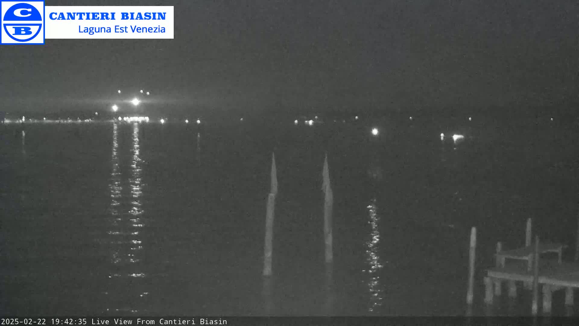 A nighttime view of a calm body of water with lights visible on the far shore and two tall posts in the foreground.