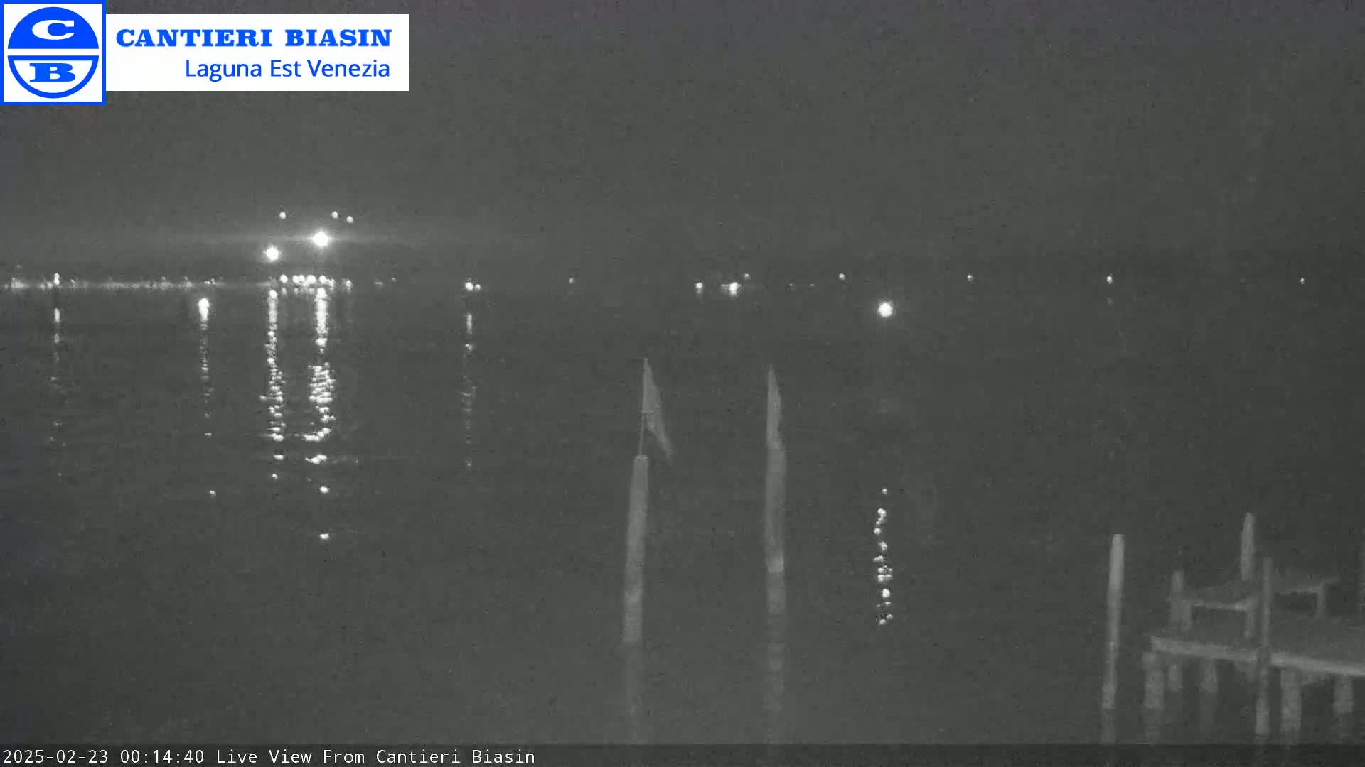 A nighttime view of calm water reflecting distant lights, with two tall posts and a small dock visible in the foreground.