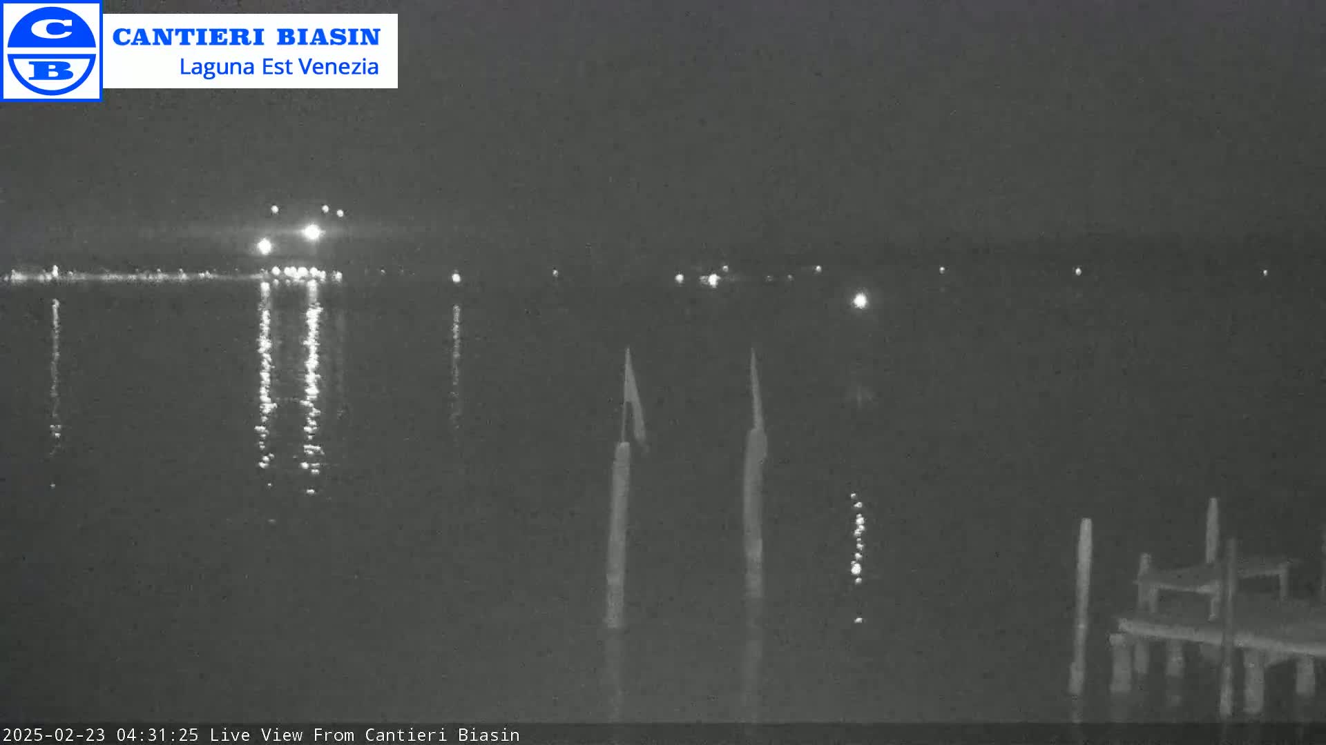 A nighttime view of a calm body of water shows reflections of lights on the surface, with a few posts and a small dock visible in the foreground.