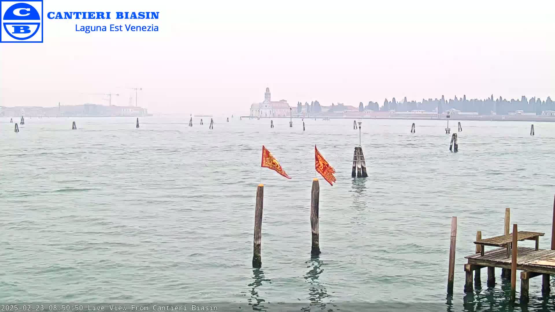 A hazy view across calm water shows a distant cityscape and two orange flags on wooden posts in the foreground, with a small wooden dock to the lower right.
