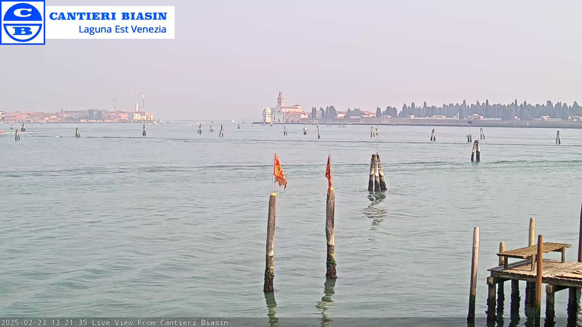 A calm body of water with wooden posts and a small dock in the foreground, and a hazy view of a distant island with buildings in the background.