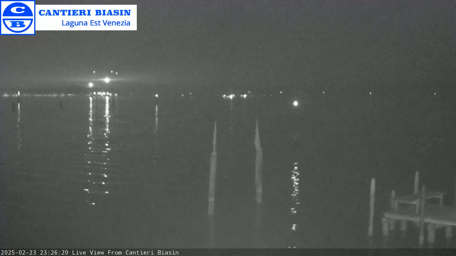 A nighttime view of a calm body of water with lights reflecting on the surface, a few distant lights visible across the water, and some dock pilings in the foreground.