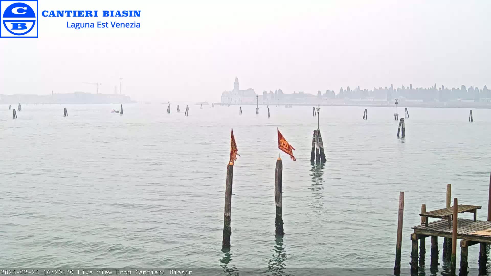A hazy, calm body of water shows a distant city skyline, several wooden posts in the water, and a small wooden dock in the foreground.