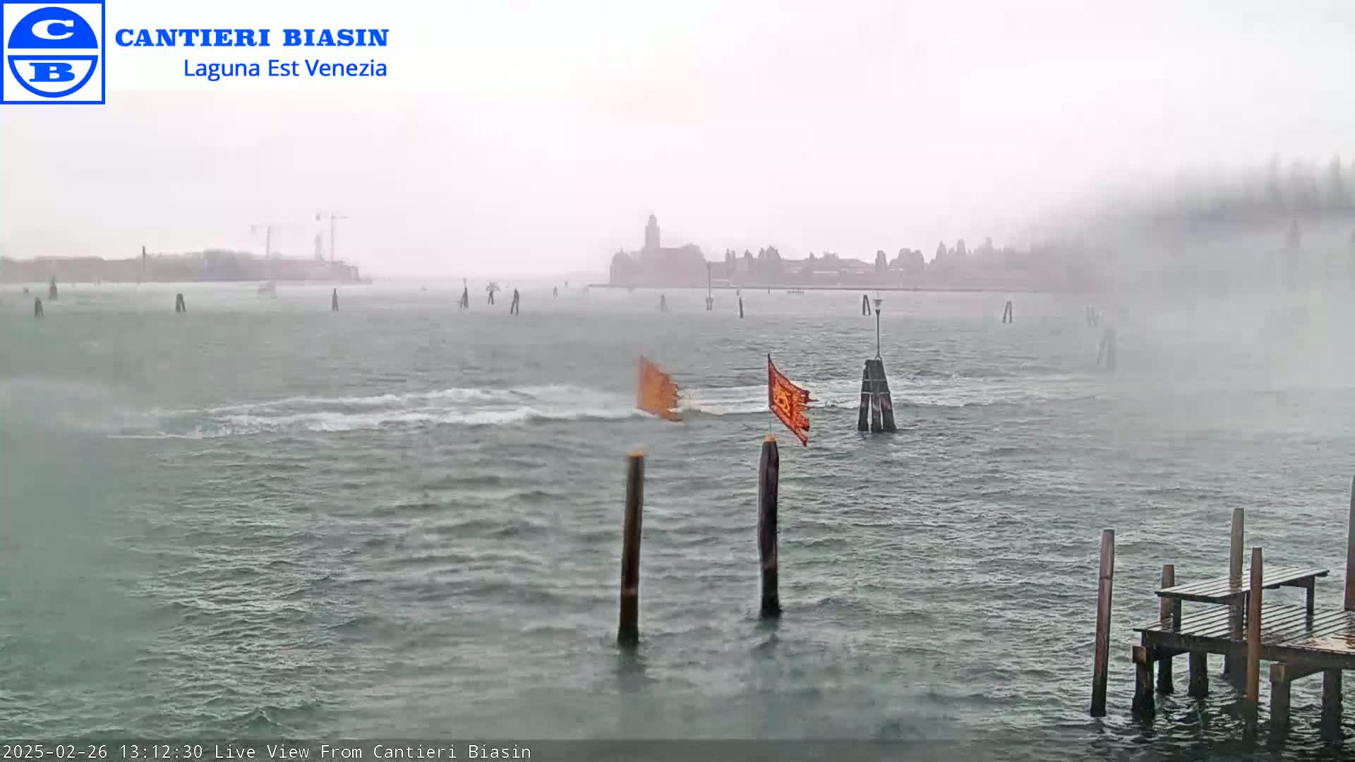 A choppy body of water with a distant city skyline under heavy fog and mist, with two orange flags affixed to posts near a small wooden dock.