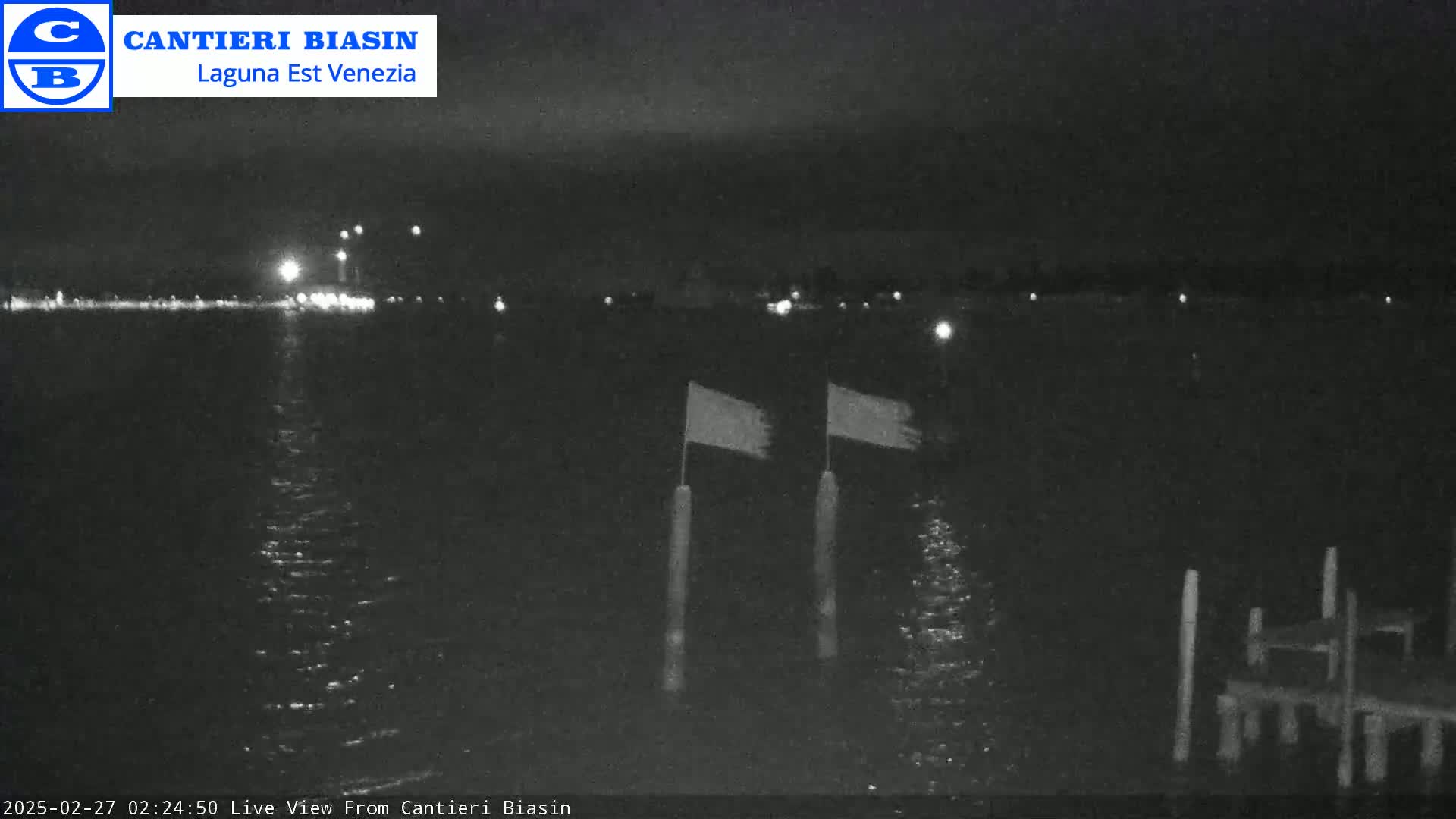 At night, a dark body of water reflects distant lights, and two flags flap on posts near a dock.