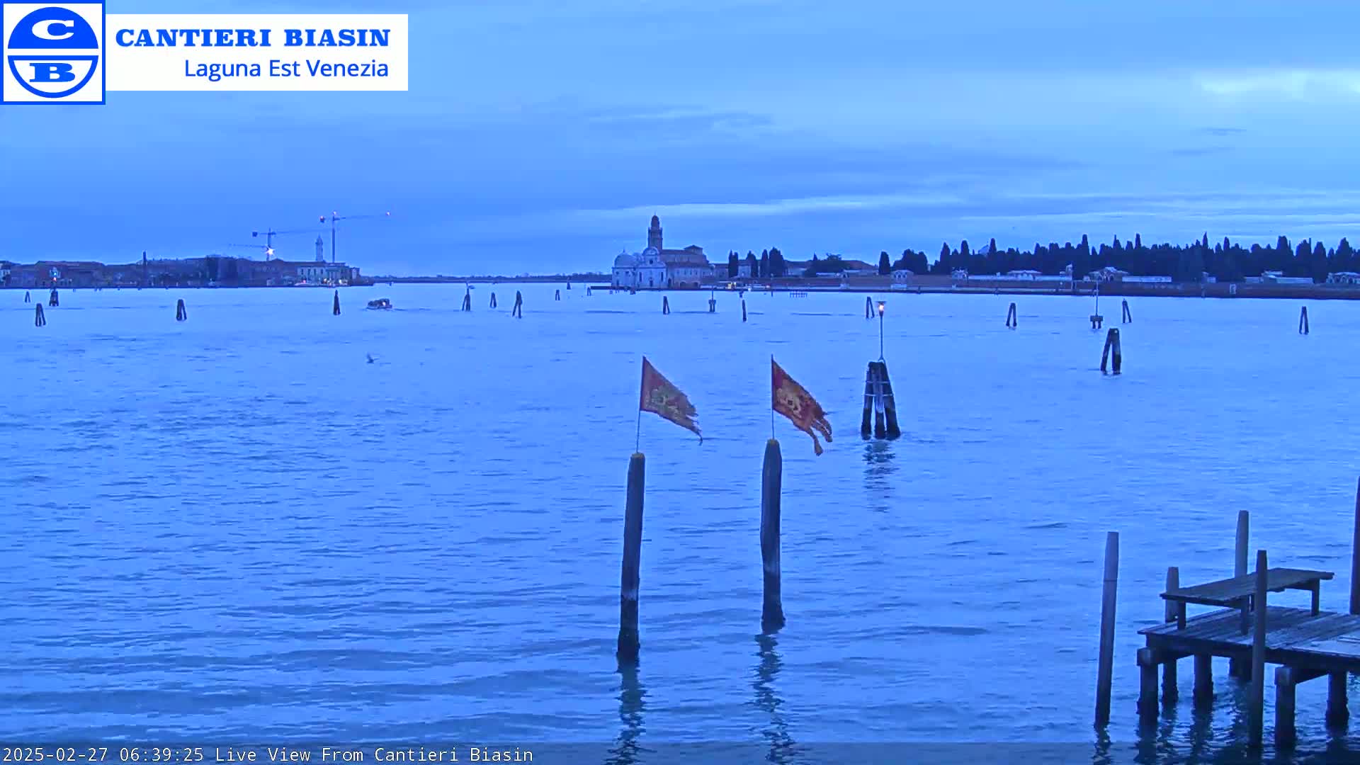 A tranquil blue body of water under a twilight sky features a distant island with buildings, several wooden posts in the water, and two flags on posts.