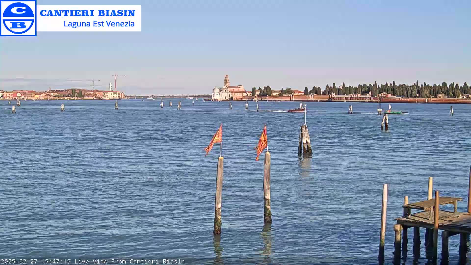 A calm body of water shows a distant island with buildings under a clear blue sky, and two orange flags are on posts in the foreground along with a small wooden dock.