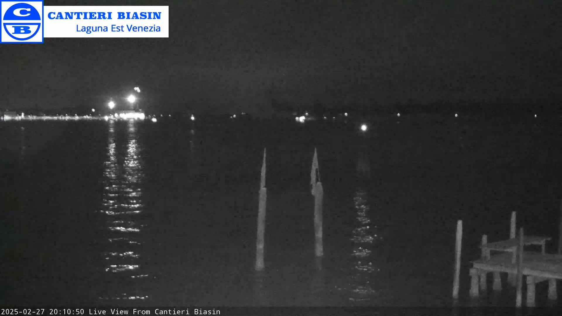 A nighttime view of calm water reflecting lights from a distant shore, with two tall posts and a small dock visible in the foreground.