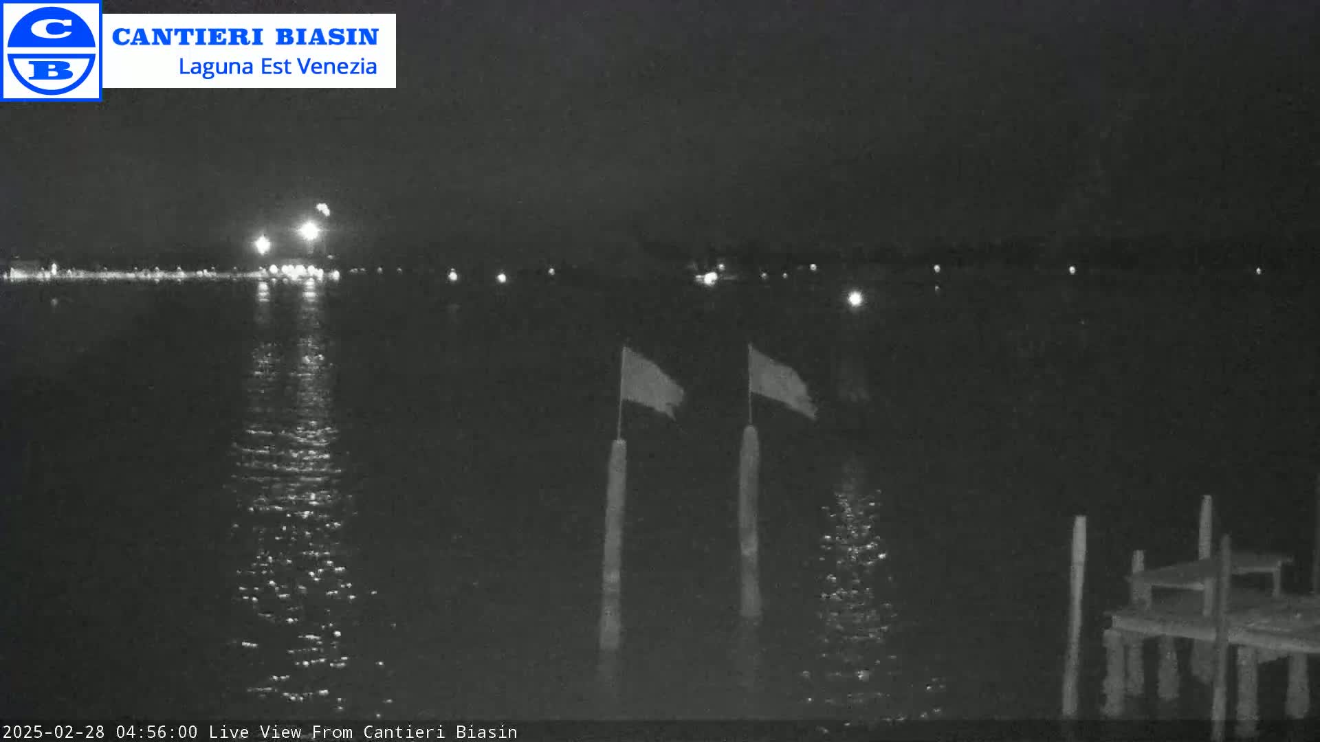 A nighttime view of calm water reflecting lights from a distant shoreline, with two flags on poles and a small dock in the foreground.