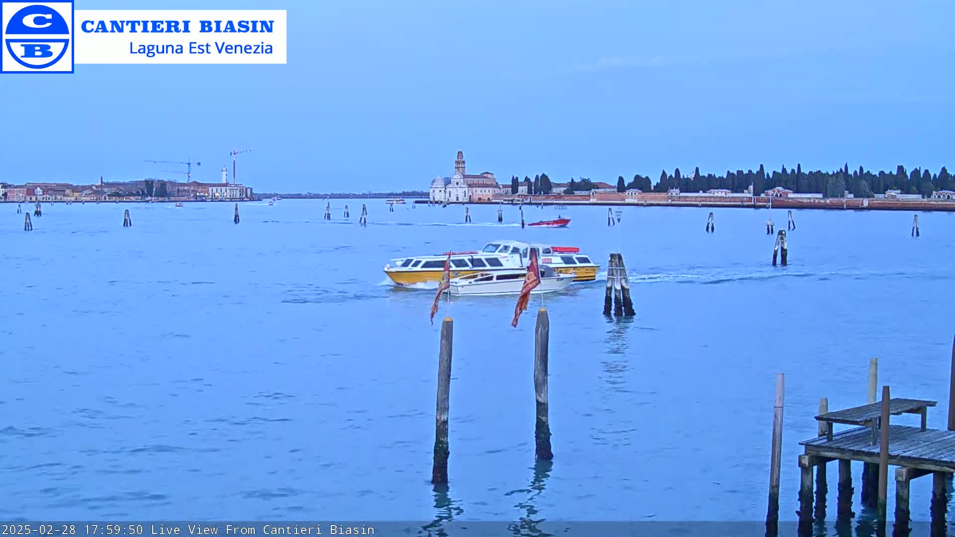 Under a clear, light-blue sky, two water taxis travel across calm water toward a distant Venetian island with buildings and trees.