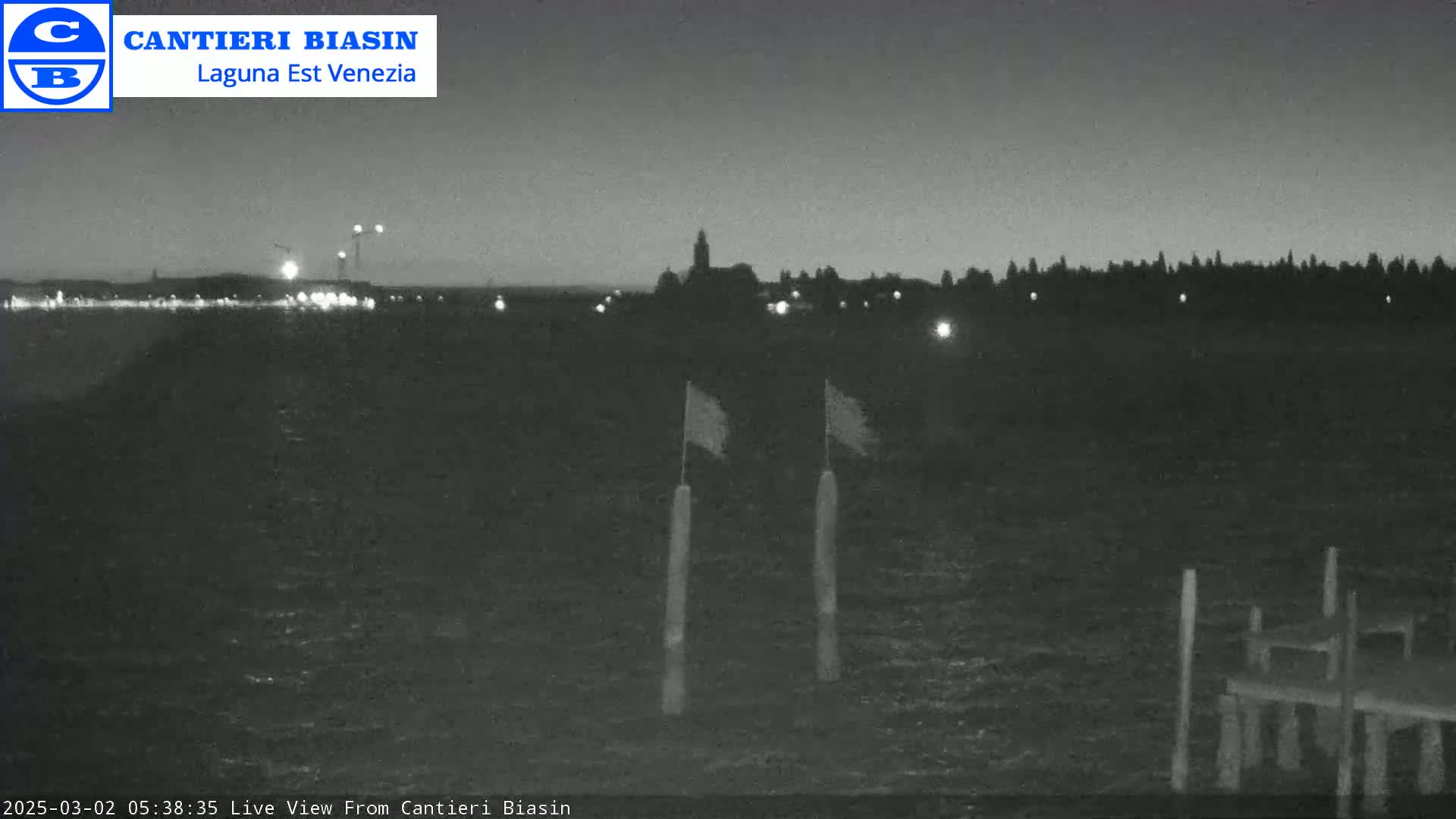 A nighttime, low-light view of a body of water shows a distant shoreline with some lights and a small dock in the foreground with two flags on posts.