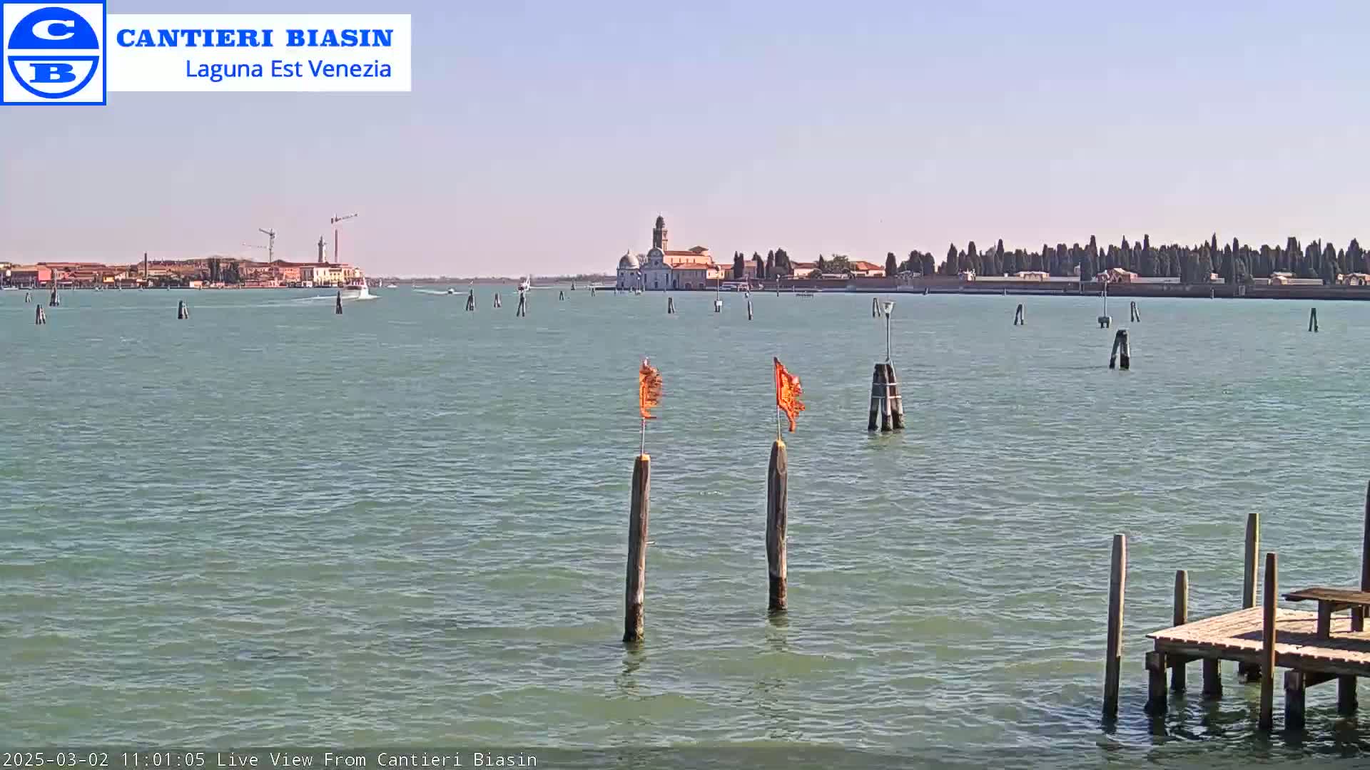 A calm, sunny day shows a body of water with several posts and orange flags, a small dock, and a distant island with buildings.