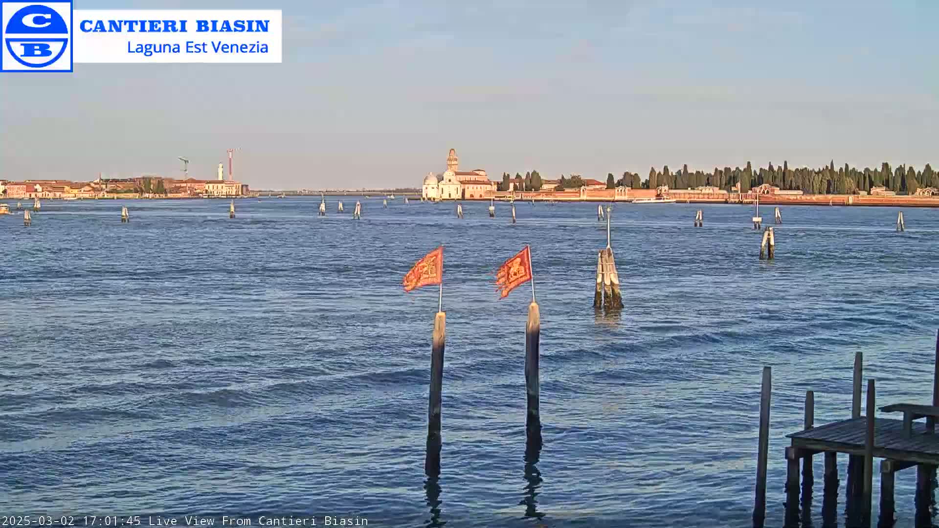 Across a calm body of water under a clear sky, two orange flags mark wooden posts in the foreground, with a distant island featuring buildings and trees visible in the background.