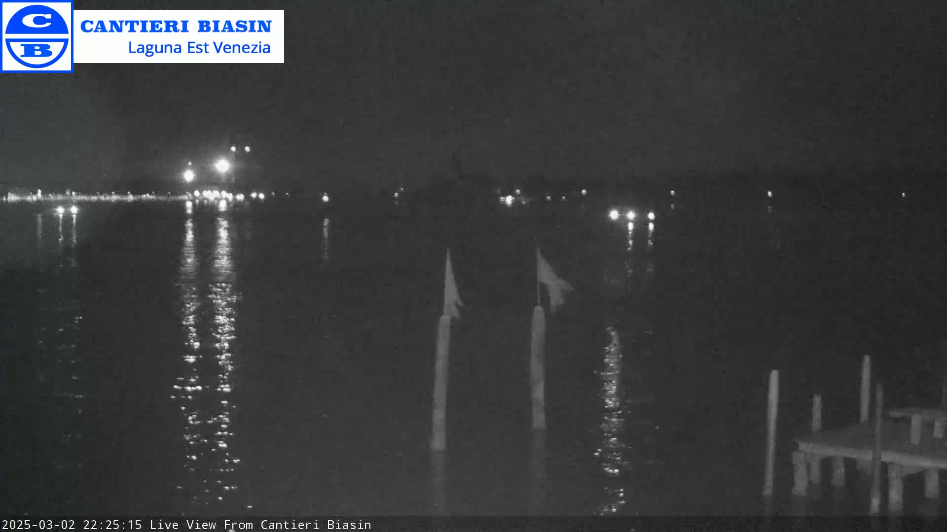 A nighttime view of calm water reflecting lights from a distant shore, with two posts and a small dock visible in the foreground.