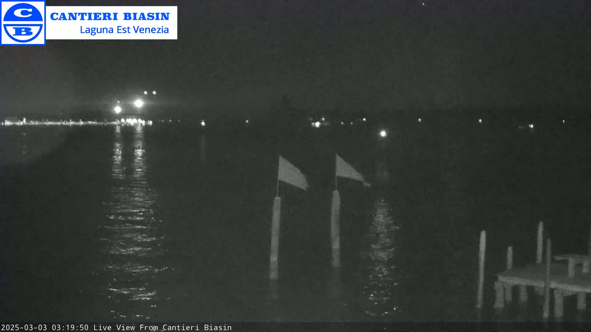 A nighttime view of calm water with two flags on posts and a small dock visible in the foreground, with lights faintly visible in the distance across the water.