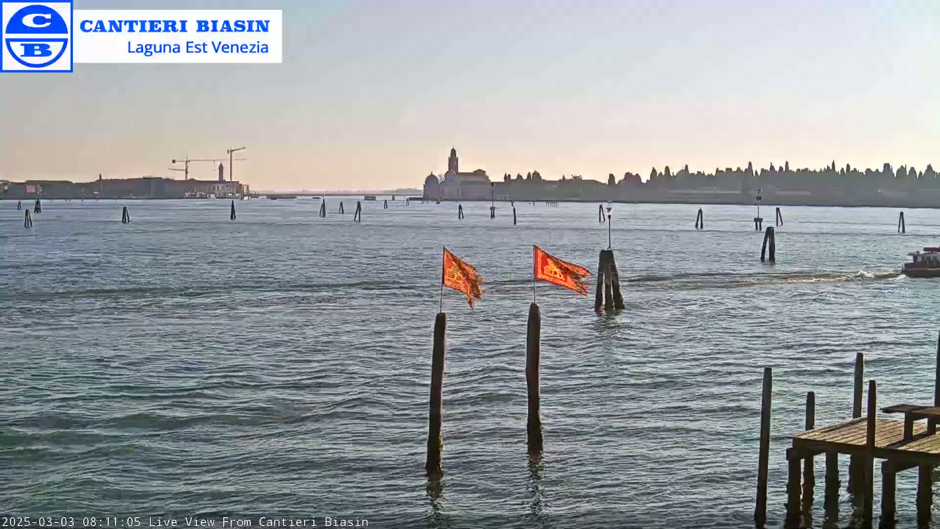 A calm body of water shows a distant island with buildings under a clear, light-blue sky, with two orange flags on wooden posts in the foreground.