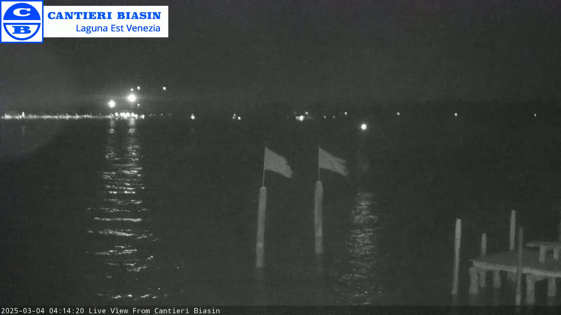 A nighttime view of calm water with two flags on poles and a small dock in the foreground, under a dark sky.