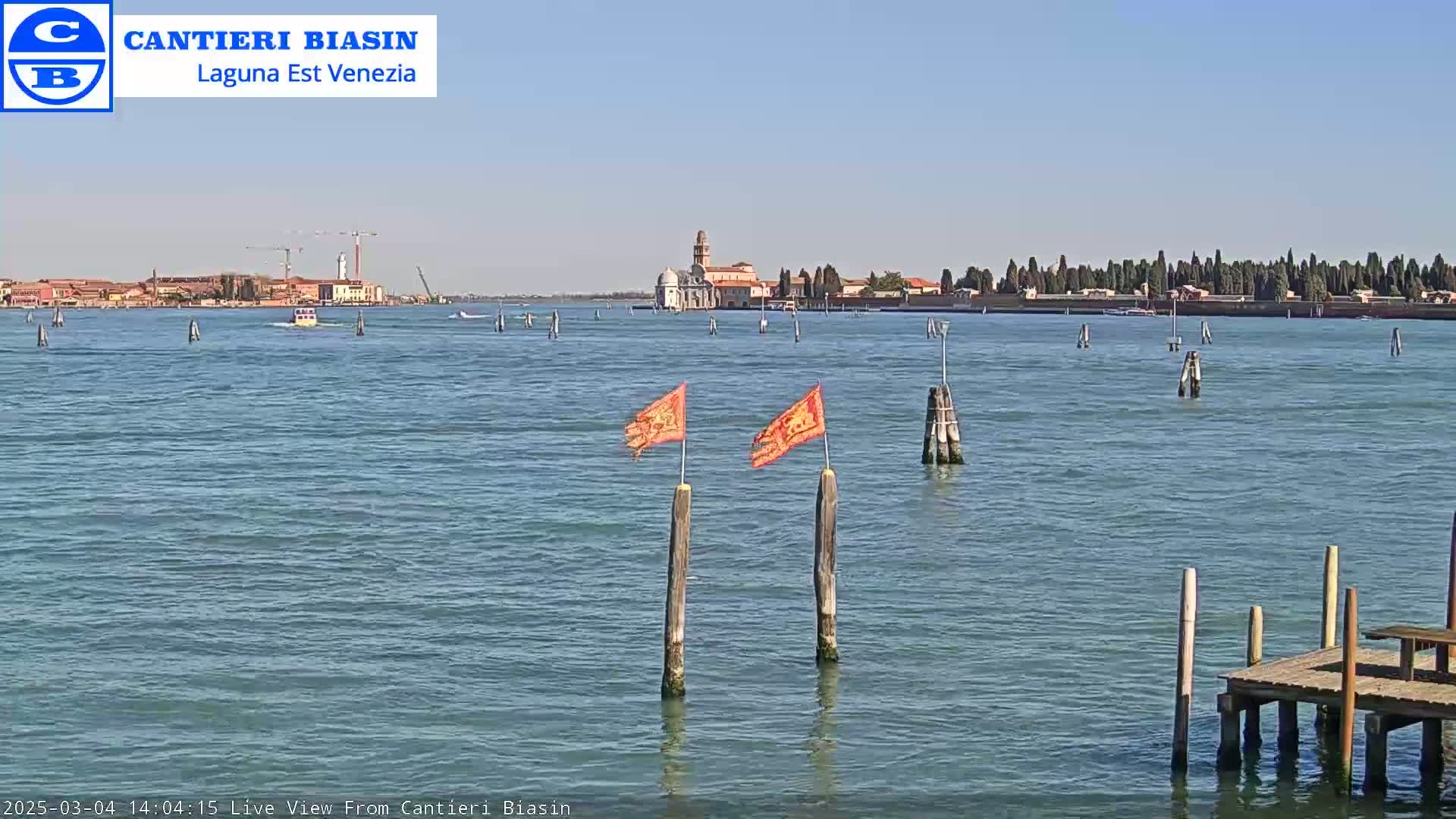 A calm body of water with a distant island showing buildings and trees under a clear blue sky, featuring two orange flags on posts in the foreground and a small dock.
