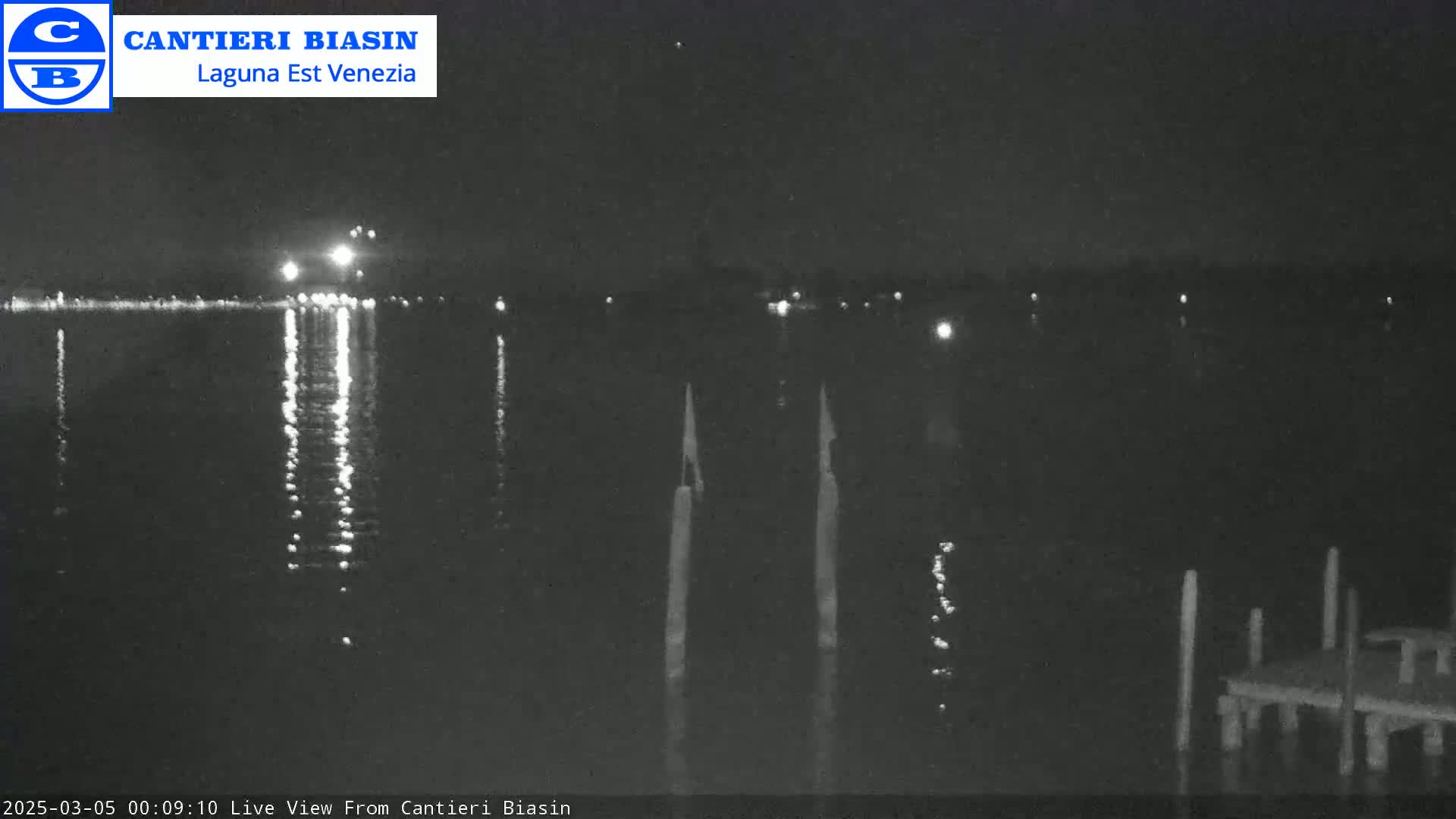 A nighttime view of a calm body of water with lights reflecting on its surface, showing a dock and pilings in the foreground under a dark sky.