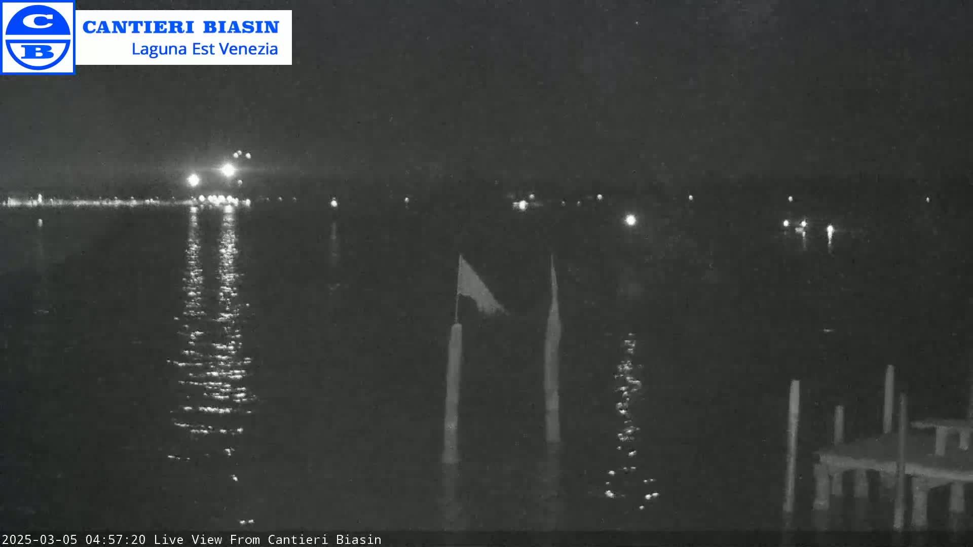 A nighttime view of a calm body of water with distant lights reflecting on the surface, a small dock, and a flagpole in the foreground.