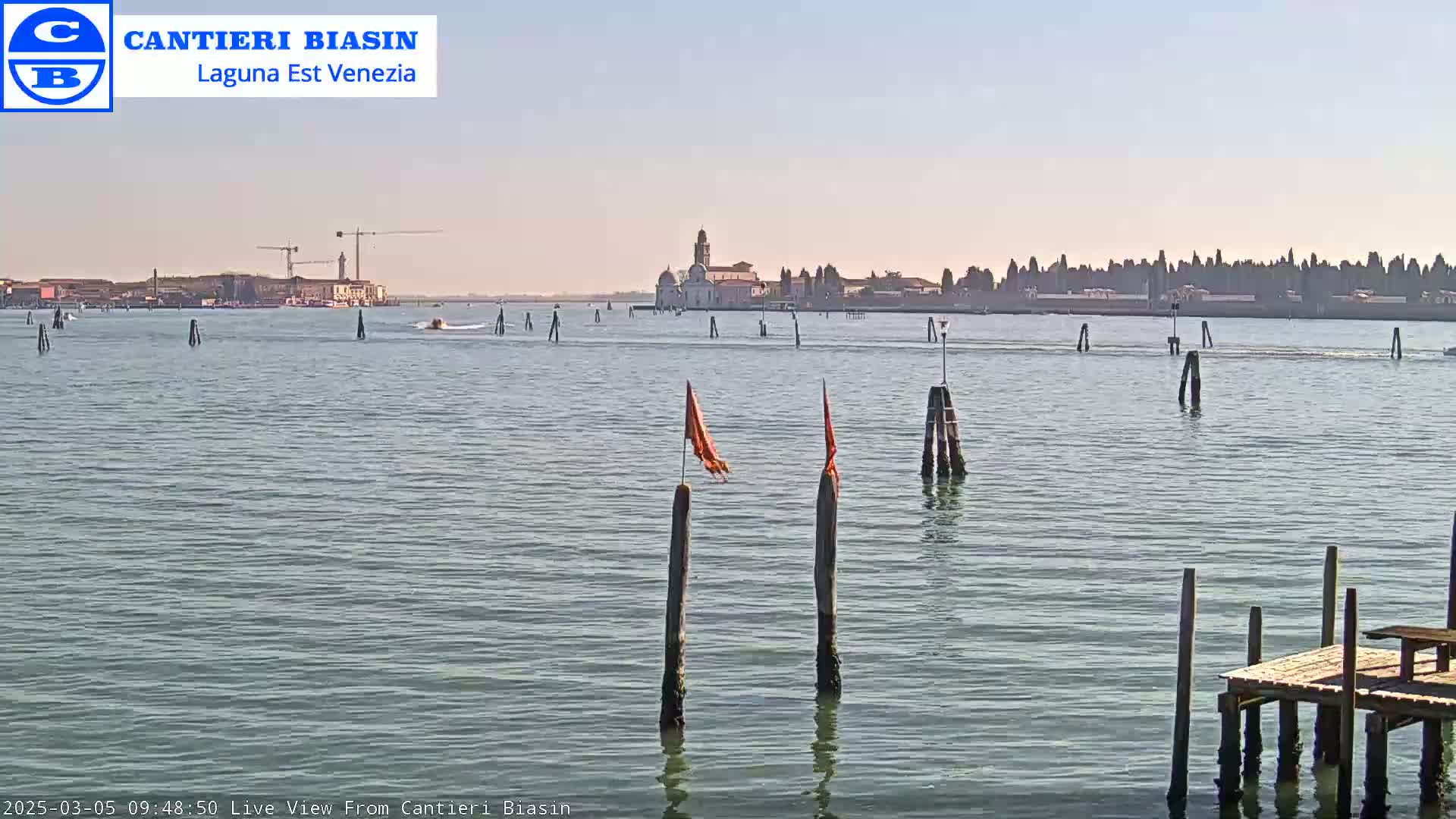 A calm body of water shows a distant island with buildings and a construction crane under a clear, sunny sky.