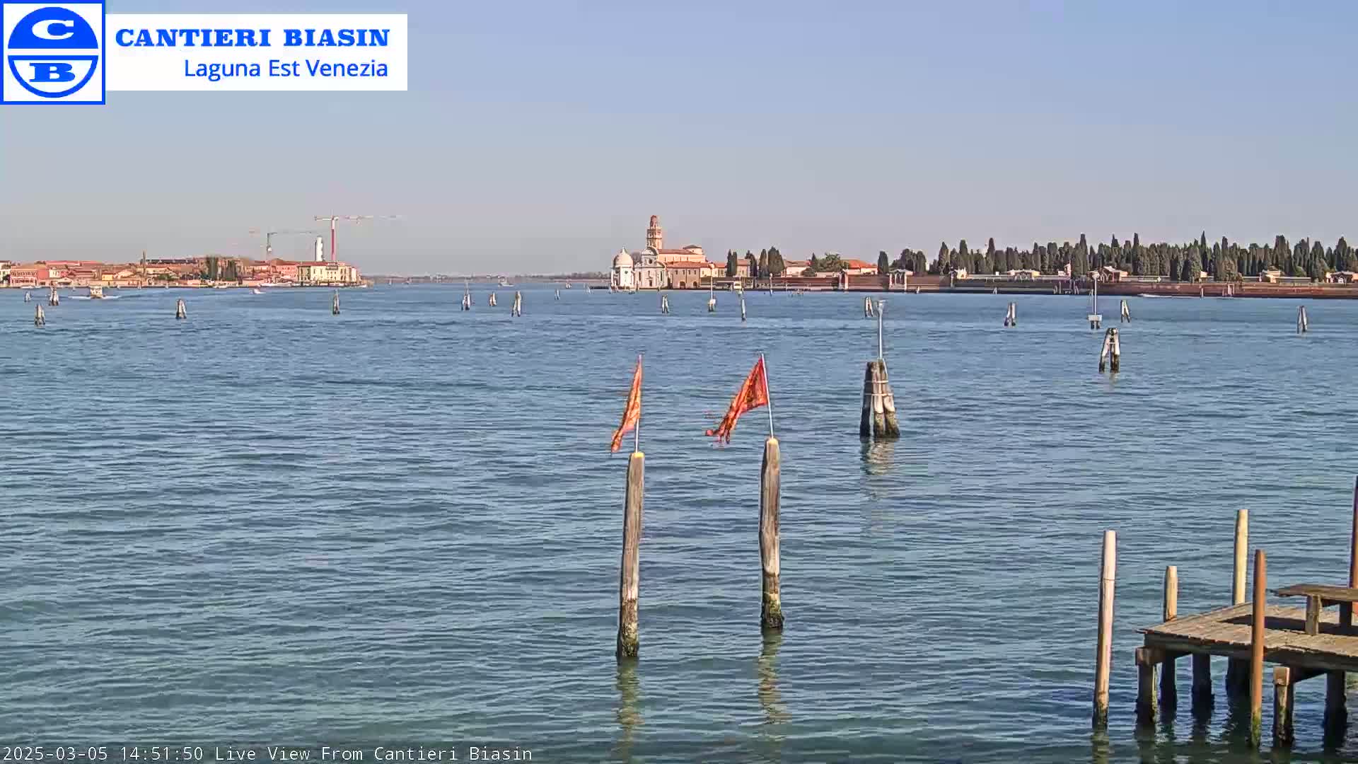 A calm body of water with a distant island featuring buildings and trees under a clear blue sky is seen from a small wooden dock.