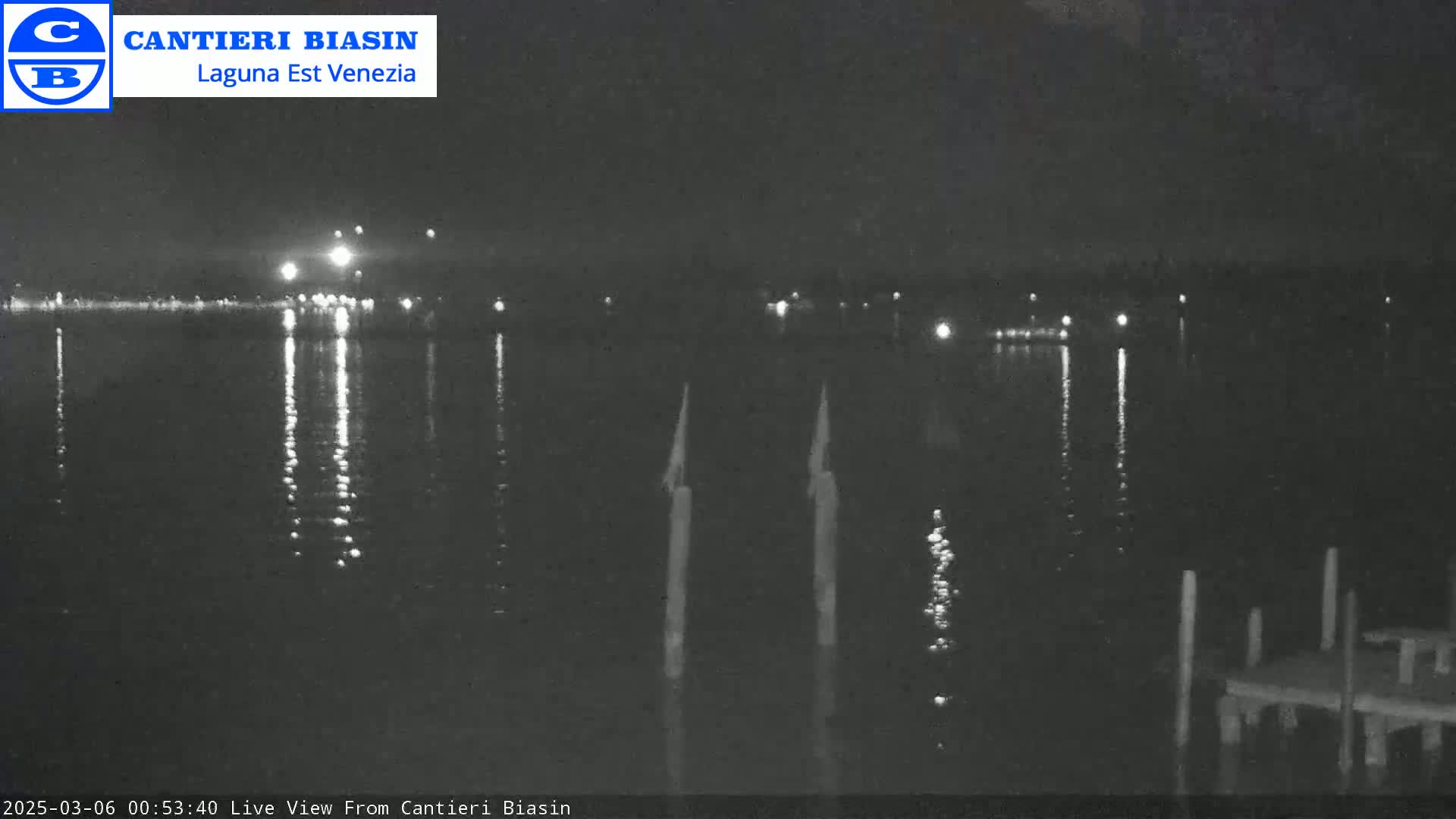 A nighttime view of calm water reflecting distant lights, with several pilings visible in the foreground.