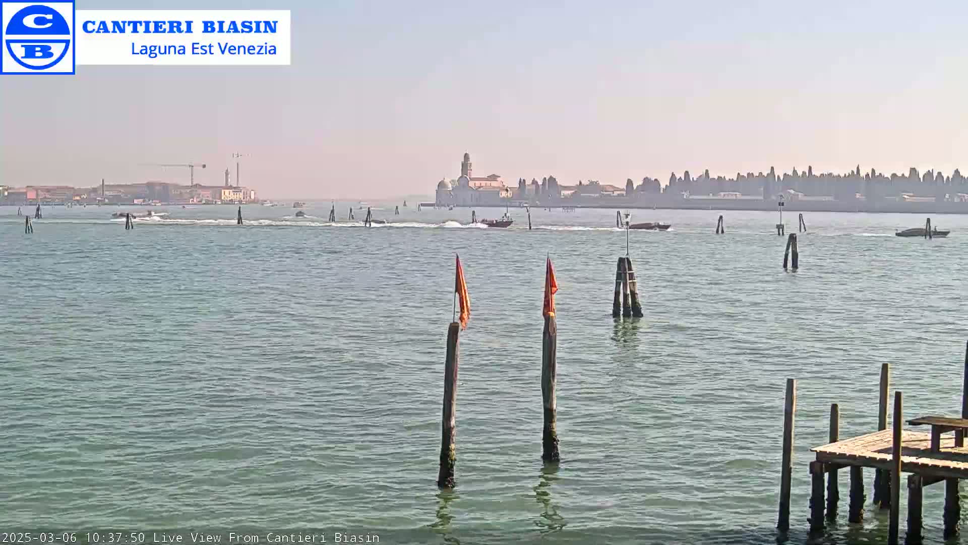 A hazy, sunny day shows a calm body of water with several boats, wooden posts, and a distant cityscape including a prominent church-like building.