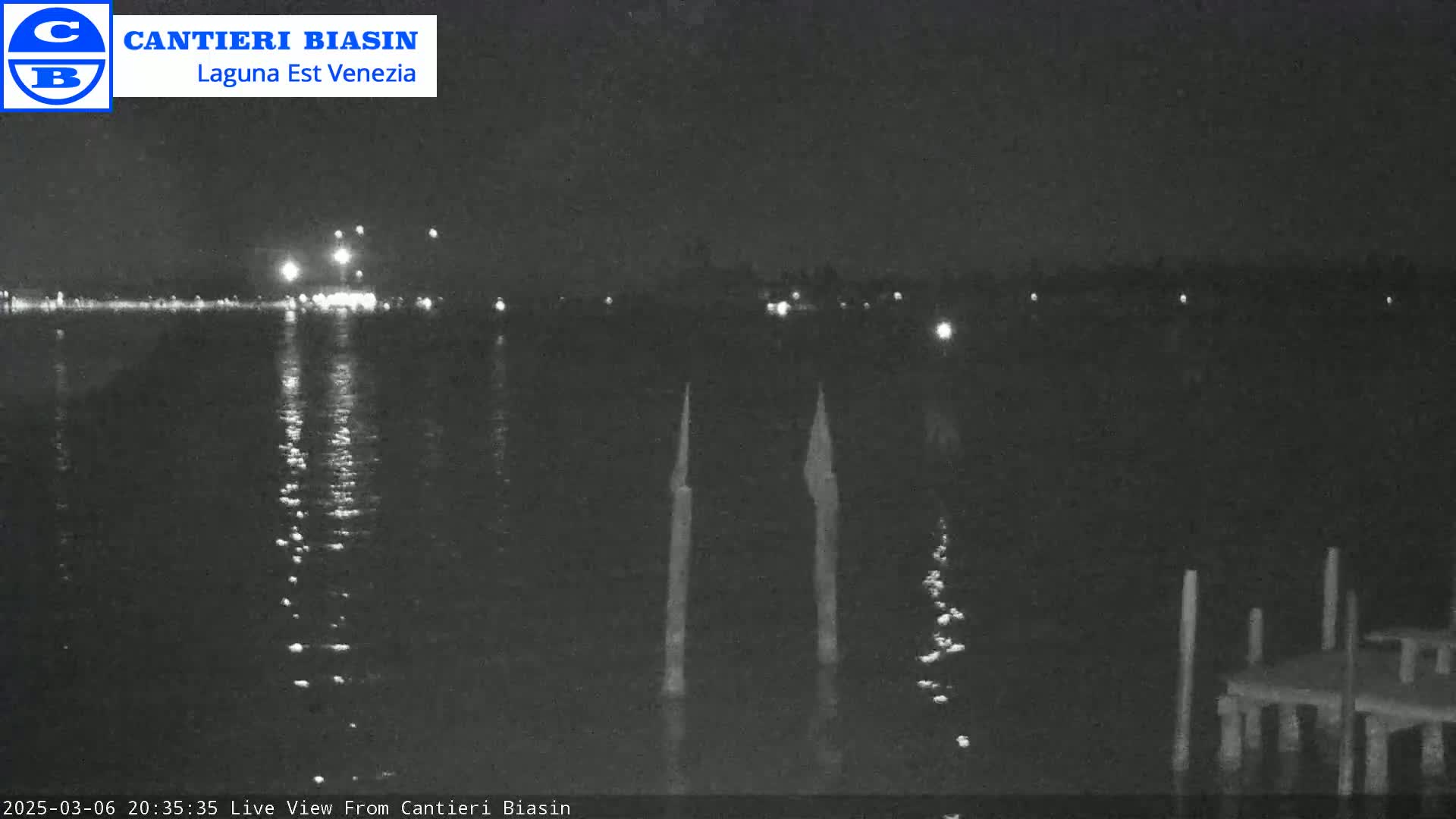 A nighttime view of calm water reflecting distant lights, with several posts visible in the foreground.